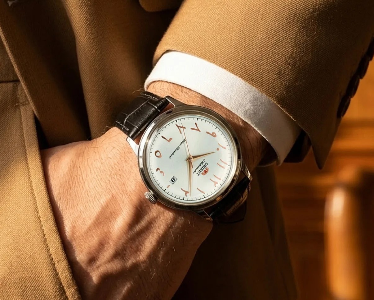 Silver wristwatch with white dial and Arabic numerals in a black leather strap worn on a wrist with a brown jacket sleeve.