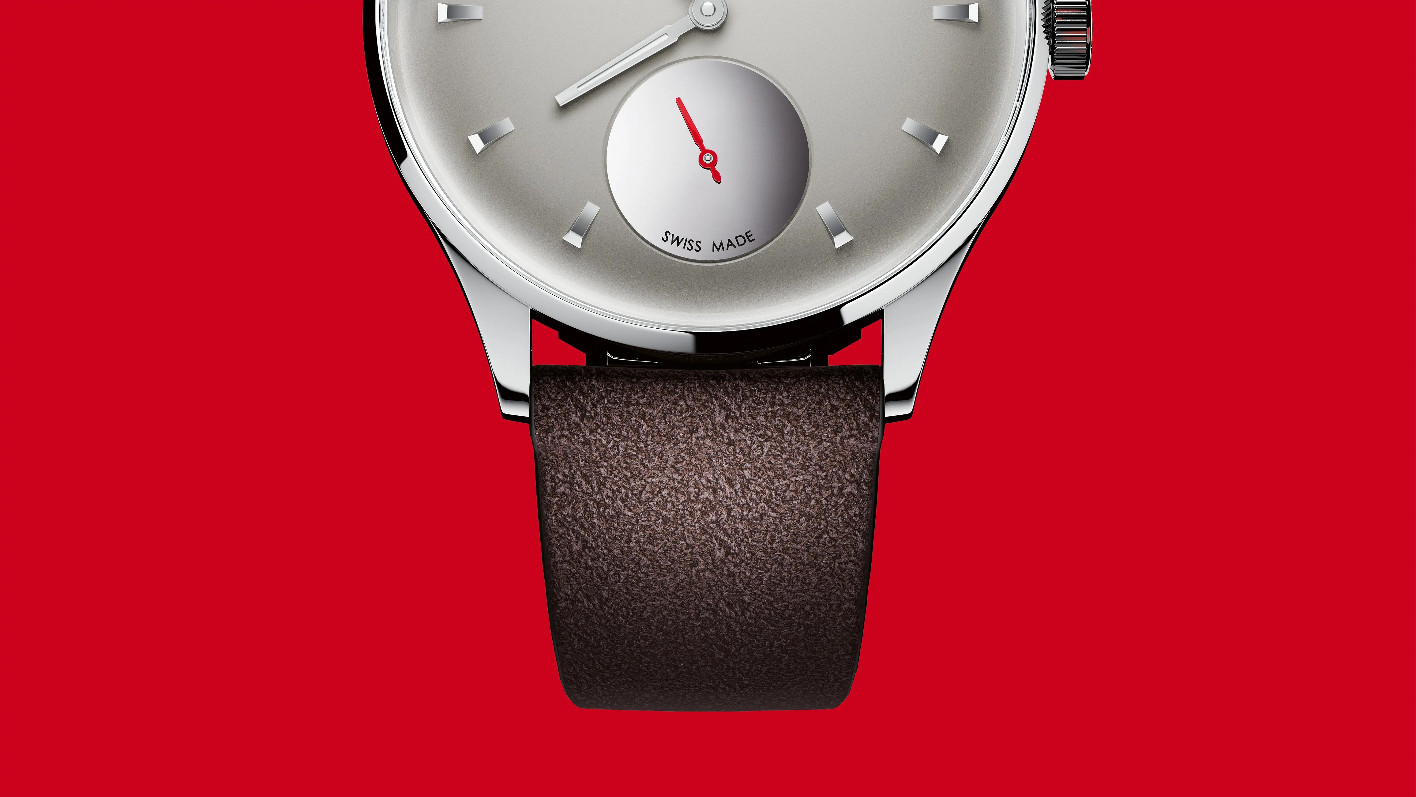 Close-up of a silver watch face with minimalist hour markers, red second hand on a subdial, and a textured brown leather strap against a red background.