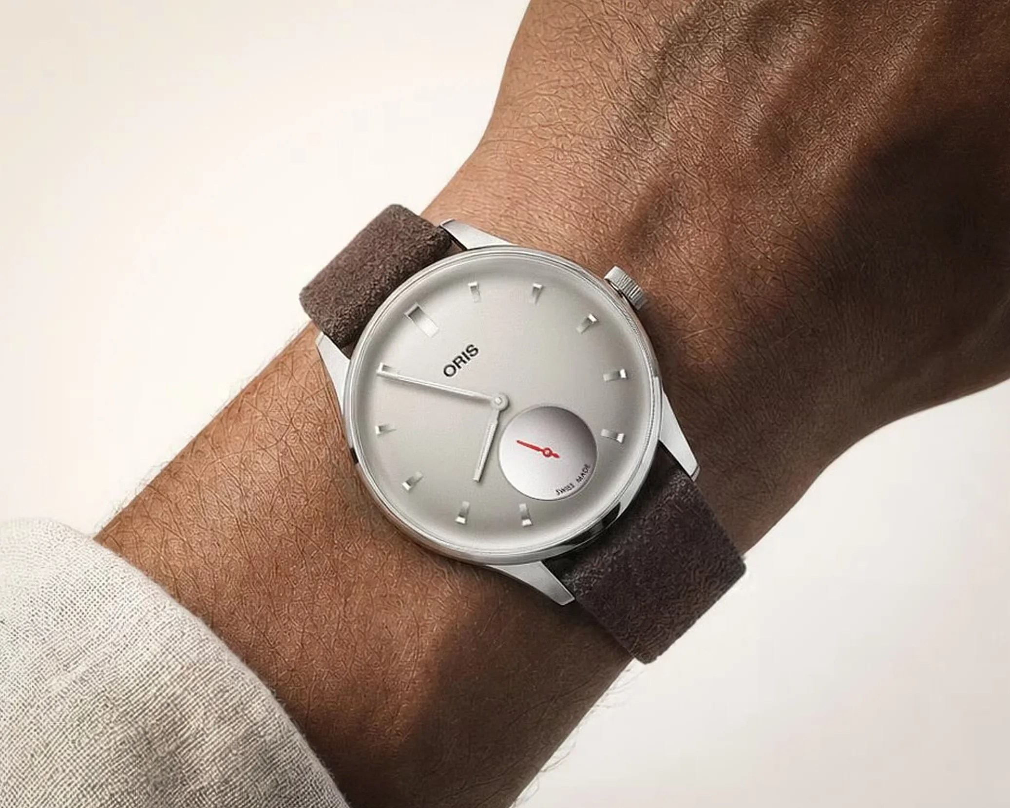 Silver Oris wristwatch with a white dial and brown suede strap worn on a wrist.