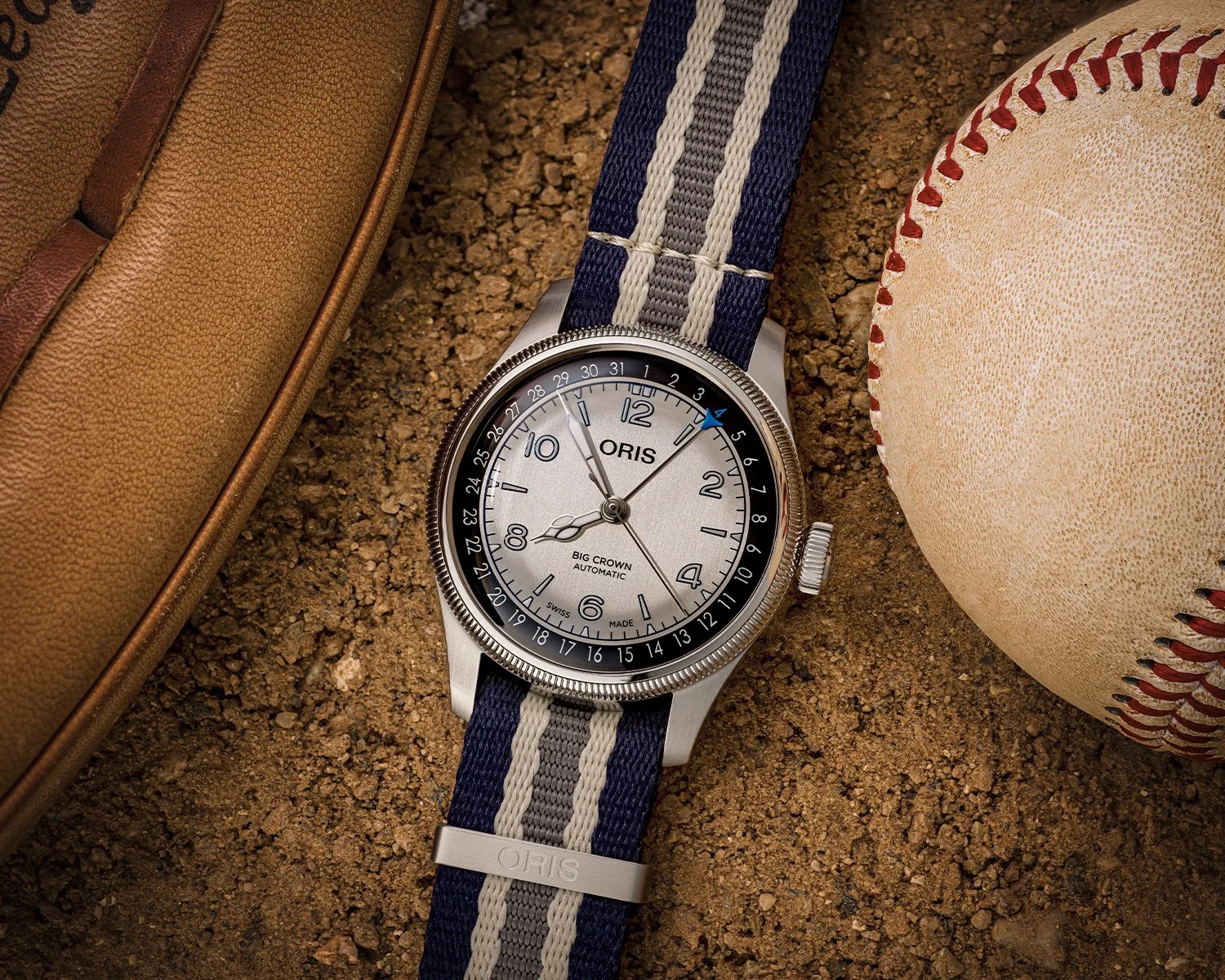 Silver Oris Big Crown automatic watch with blue and gray striped fabric strap on dirt next to a baseball and glove.
