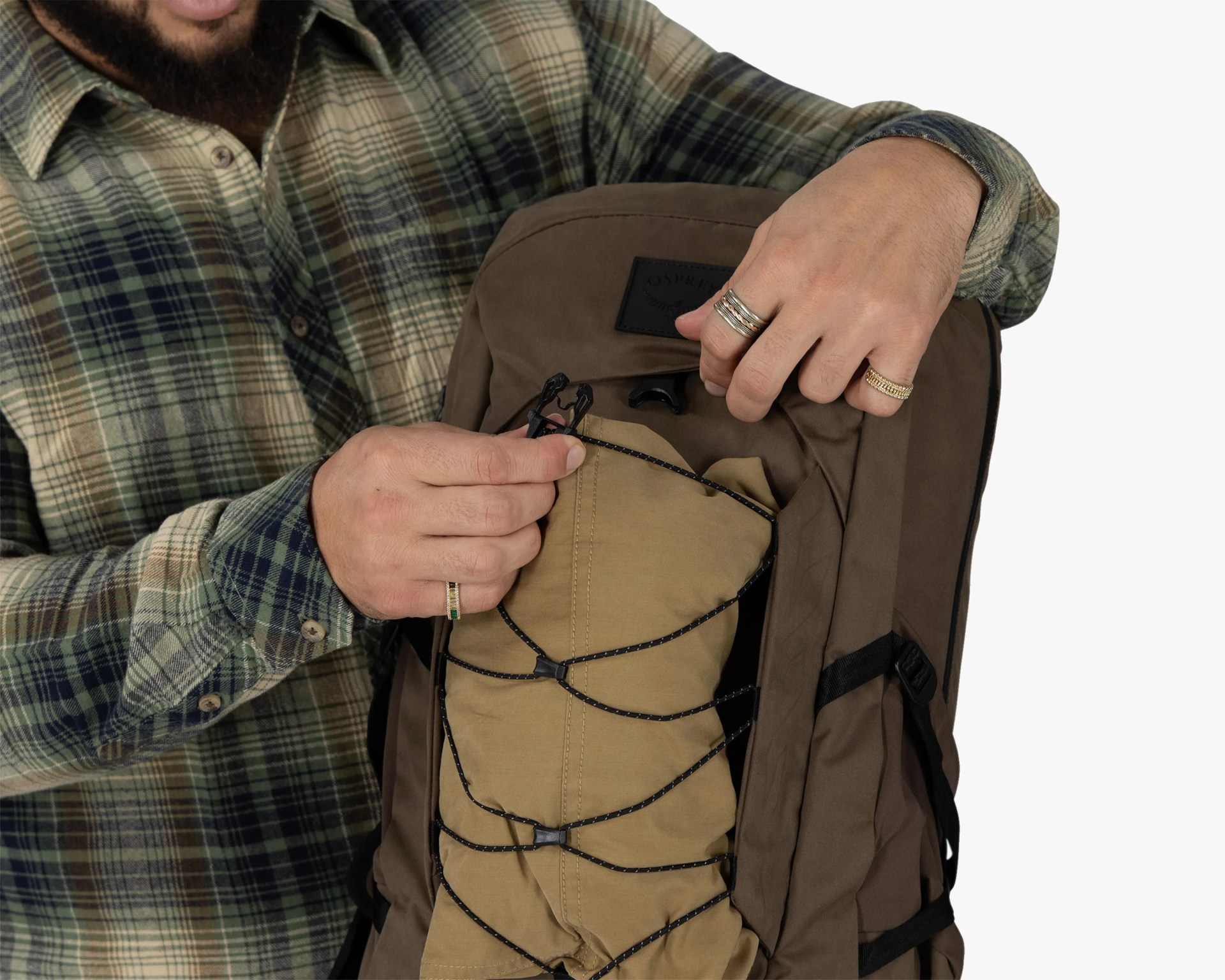 Person wearing a green and beige plaid shirt adjusting black cord on a brown Osprey backpack.
