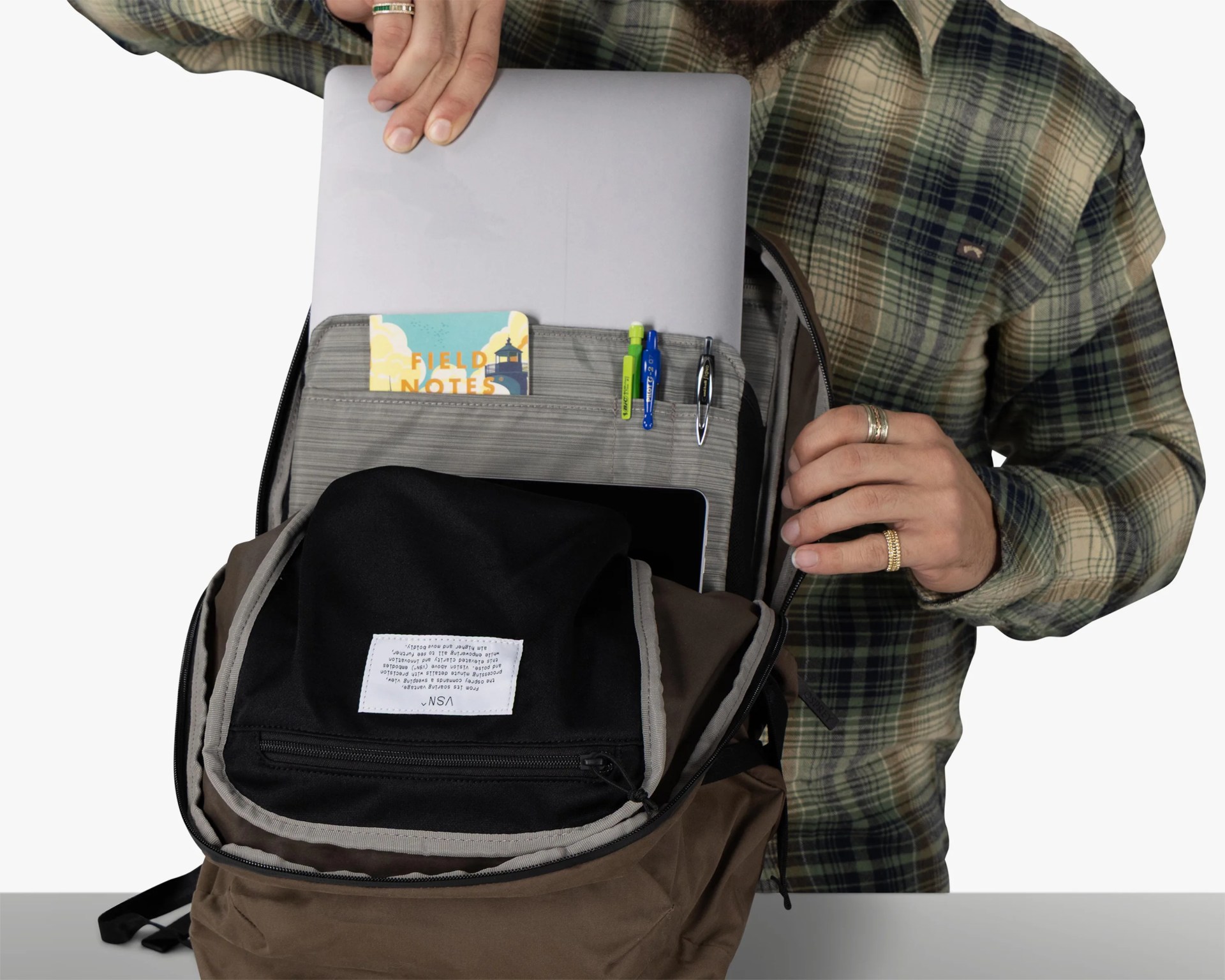 Brown backpack with a laptop, Field Notes notebook, and three pens in organizer pockets, worn by a person in a green plaid shirt.