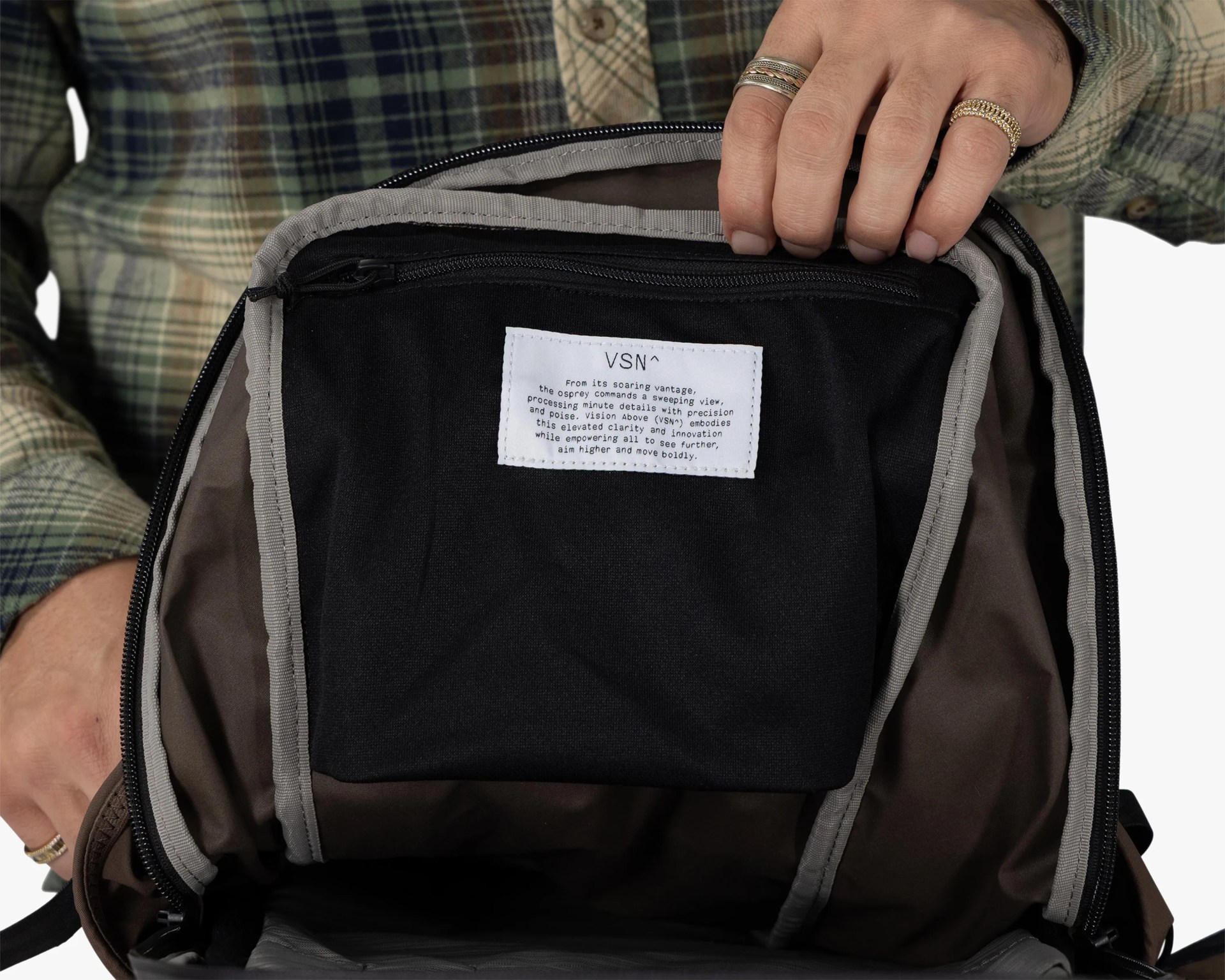 Black fabric interior pocket with a white label inside a brown backpack held open by a person wearing a green plaid shirt.