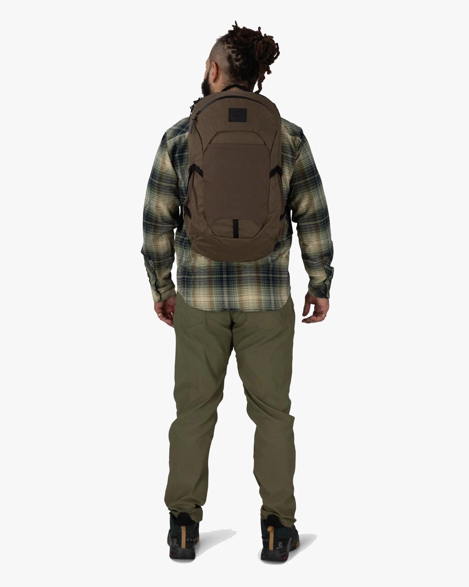 Man wearing olive green pants, a green plaid shirt, black hiking shoes, and a brown backpack, shown from the back.