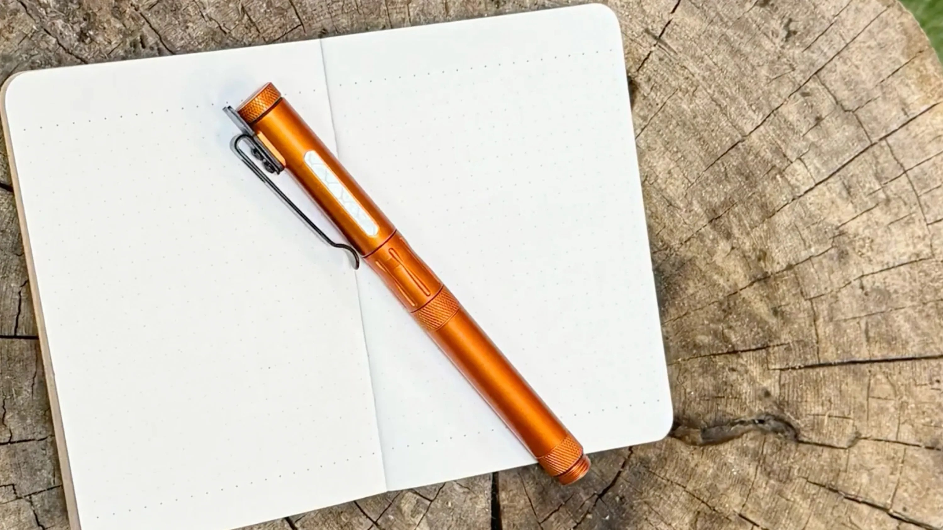 Orange metal flashlight with black clip resting on an open dotted notebook on a tree stump.