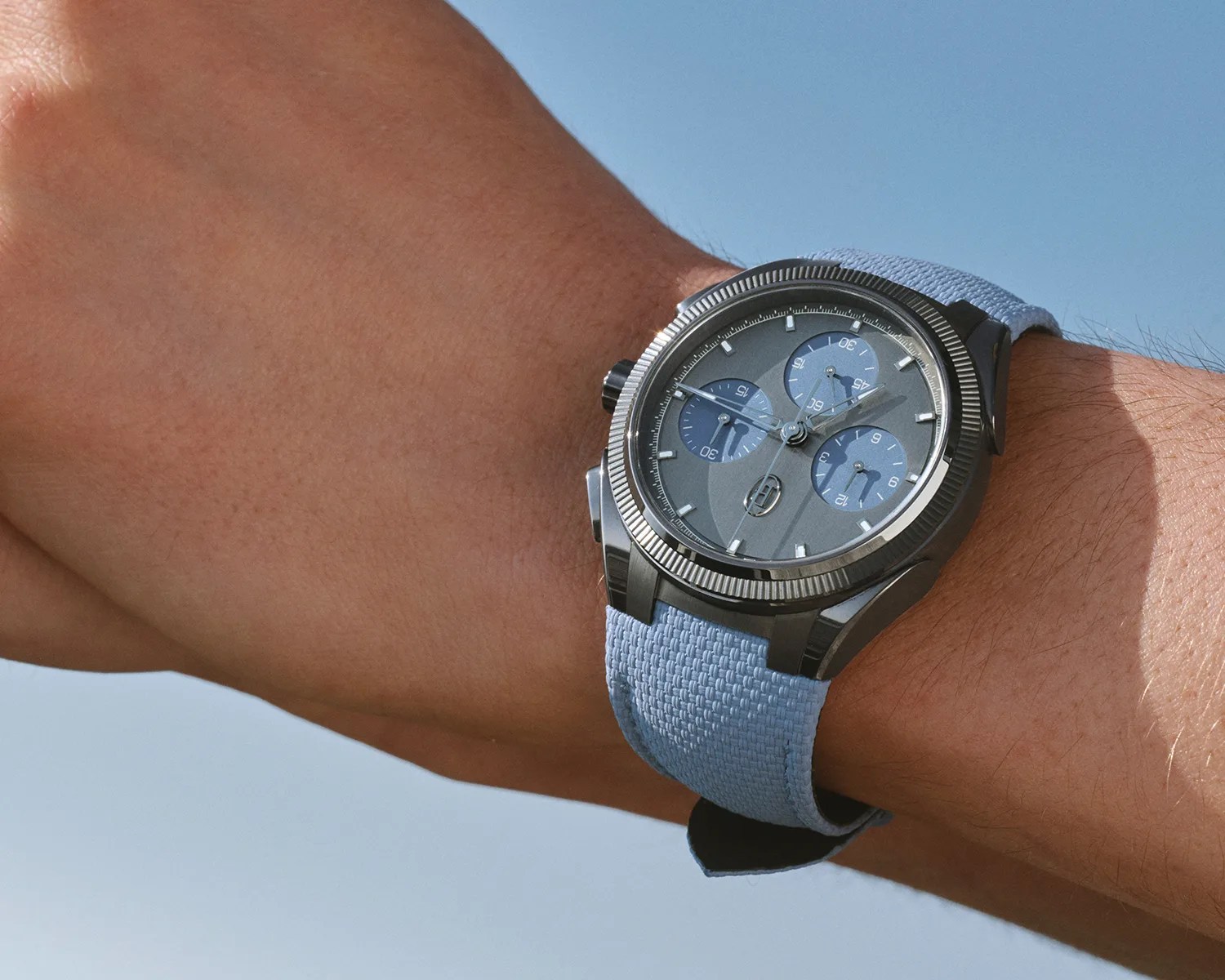 Black metal wristwatch with three blue subdials and a light blue woven fabric strap worn on a wrist.
