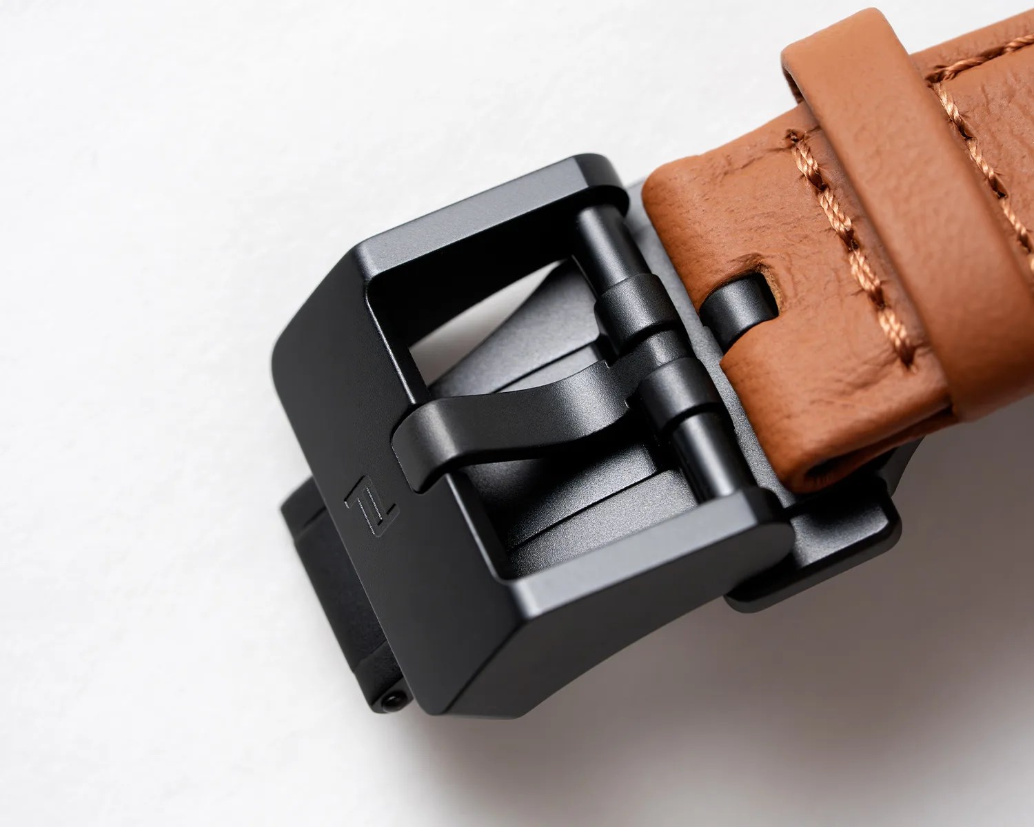 Close-up of a matte black metal buckle attached to a tan leather strap with visible stitching.