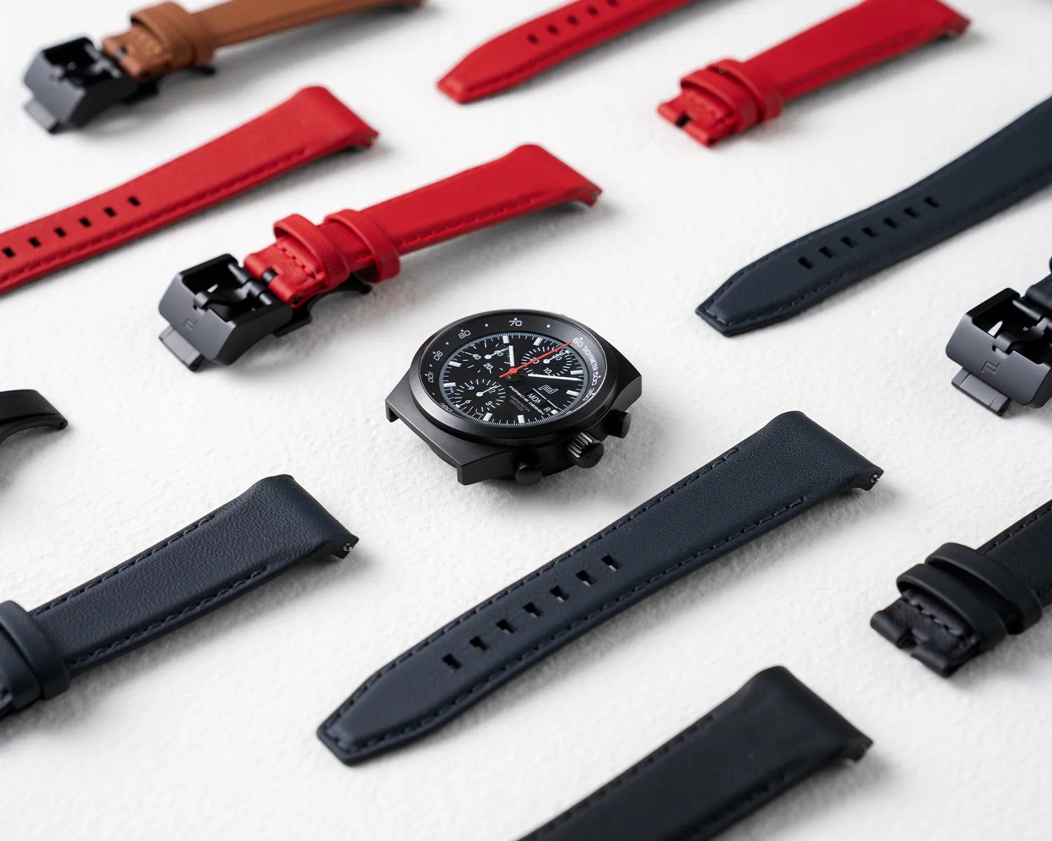 Black watch face with red second hand surrounded by multiple red and black leather watch straps on a white surface.