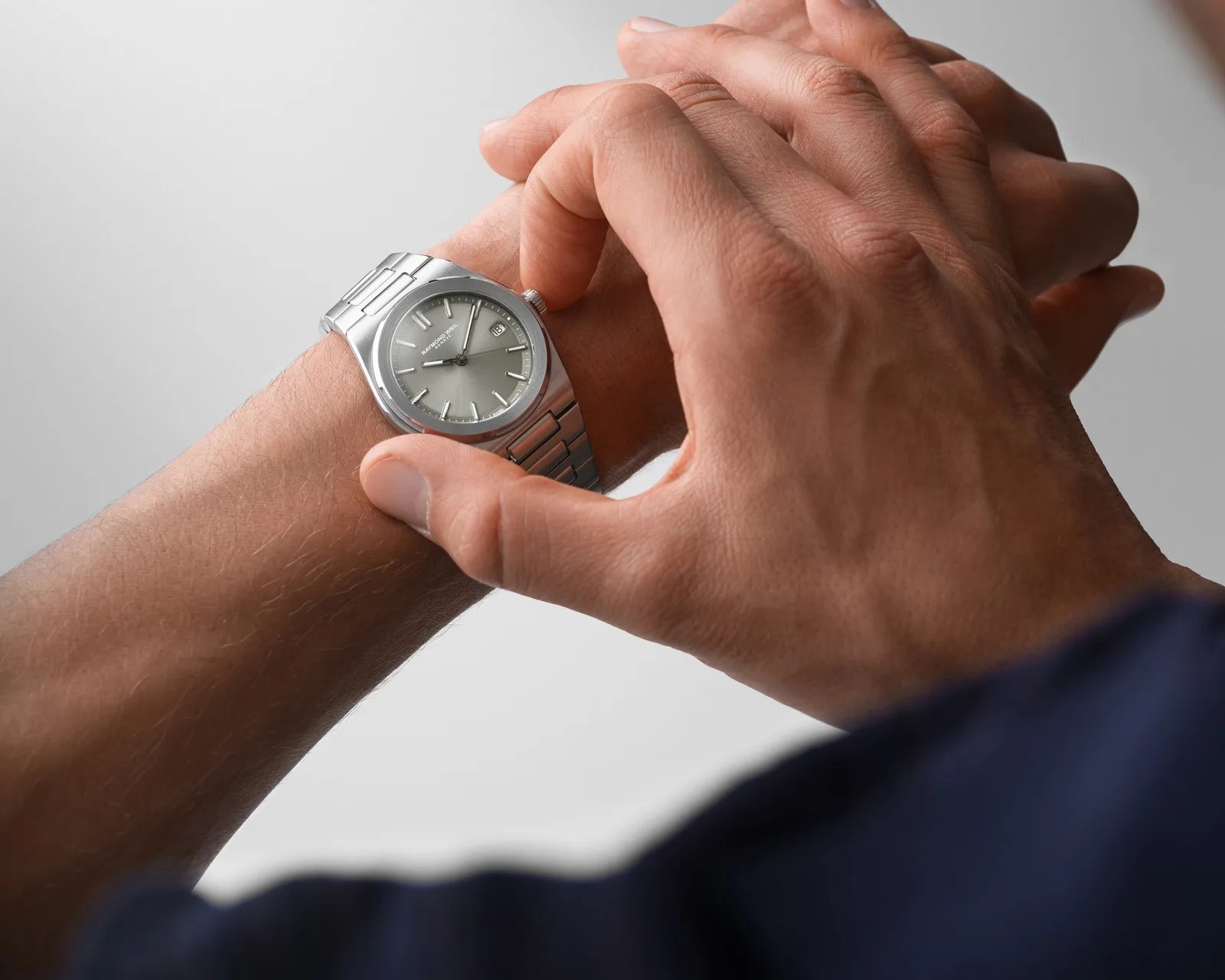 Silver Raymond Weil wristwatch with a metal link band worn on a wrist, being adjusted by a hand.