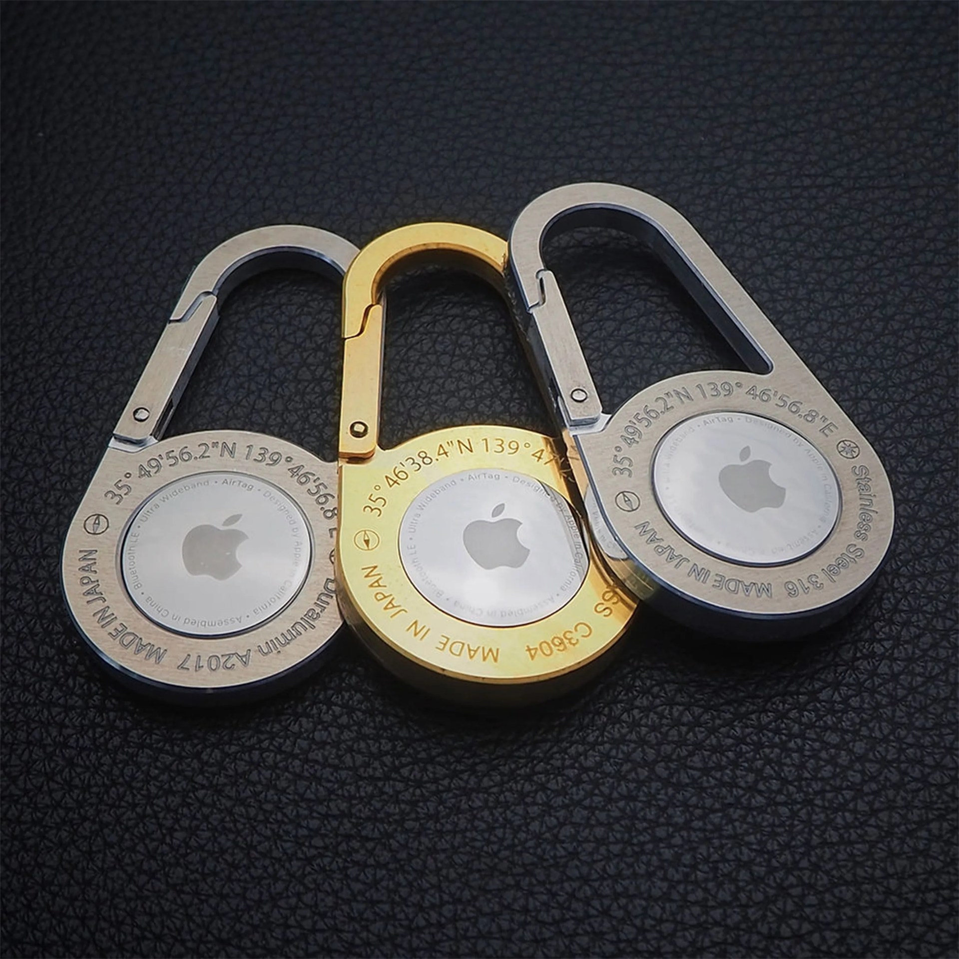 Three Apple AirTags attached to metal carabiner keychains in silver, gold, and silver colors on a black textured surface.
