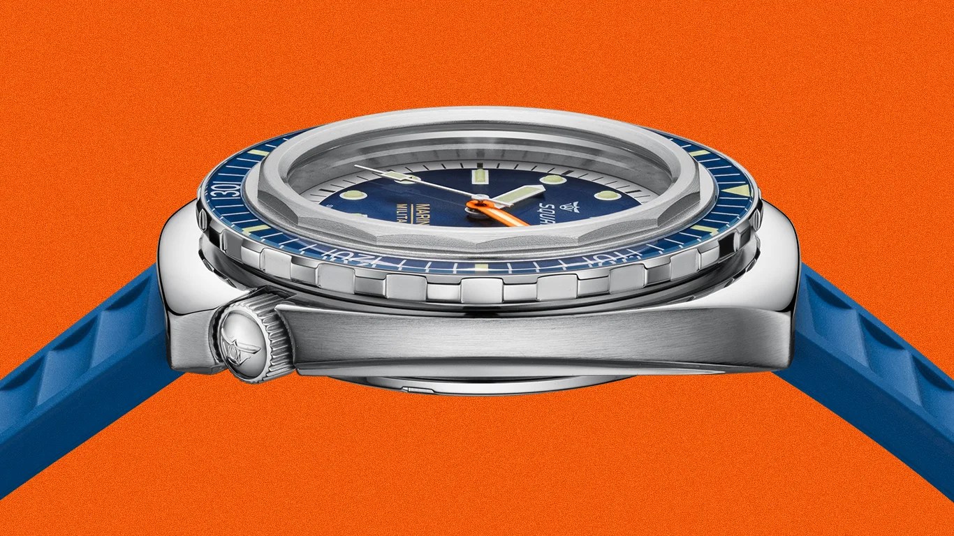 Side view of a stainless steel dive watch with a blue bezel and blue rubber strap against an orange background.