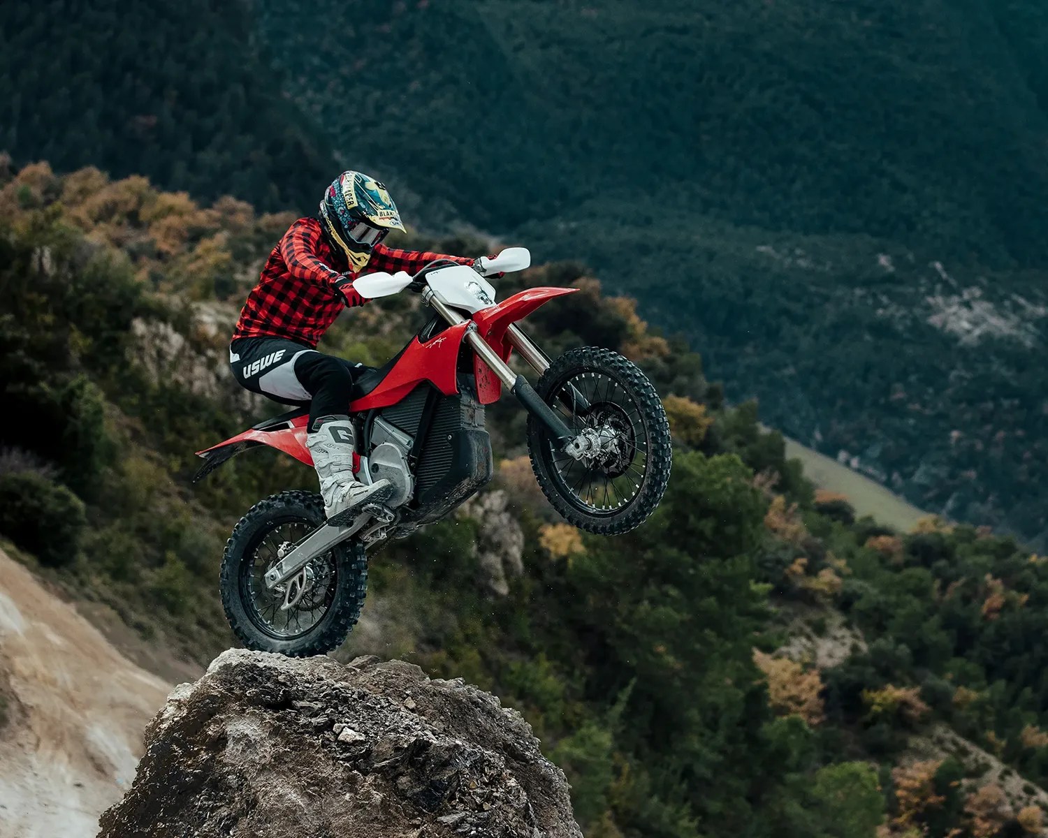 Person in a red and black checkered jacket riding a red dirt bike on a rocky terrain with forested hills in the background.