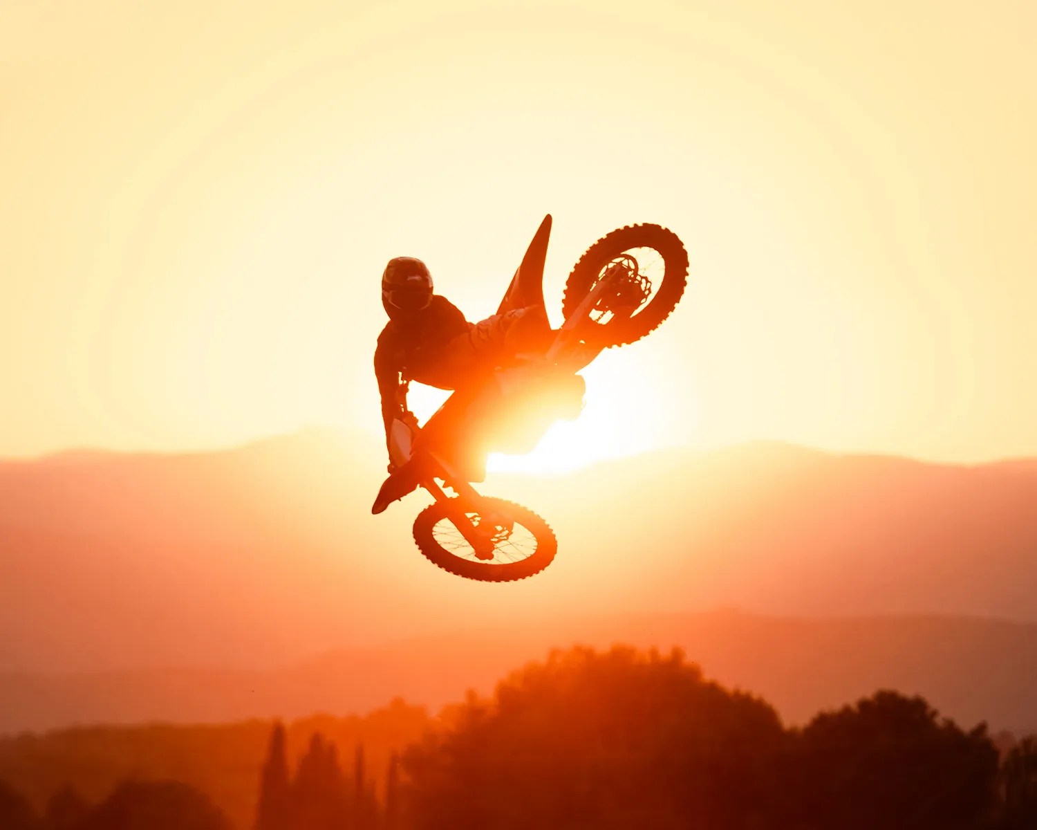Silhouetted motocross rider performing a jump against a bright orange sunset with distant hills.