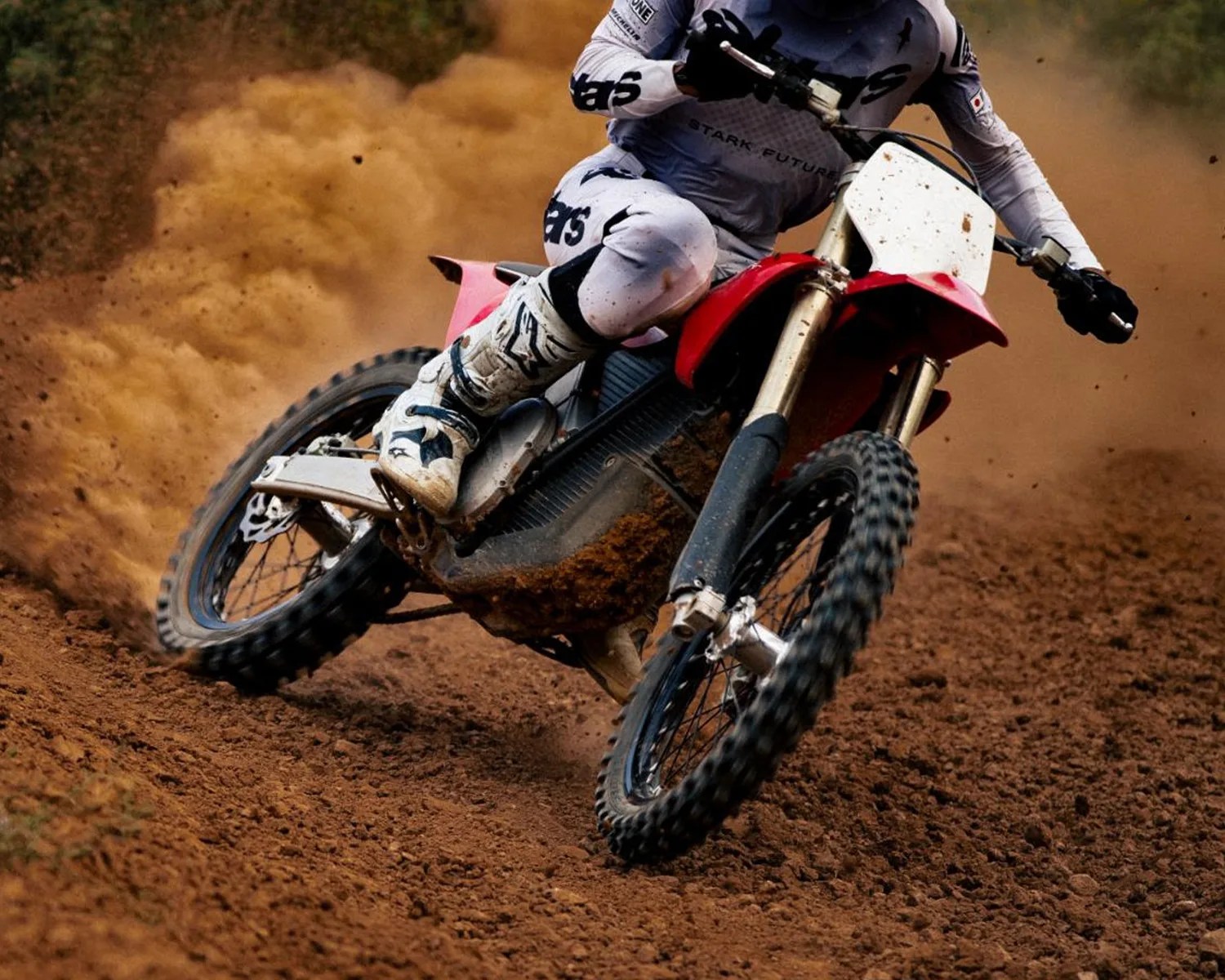 Dirt bike with red fenders kicking up dust on a dirt track, ridden by a person in white gear and boots.