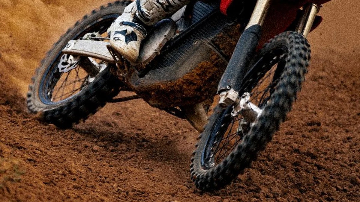 Close-up of a dirt bike with knobby tires kicking up dirt on a muddy track, rider wearing white boots.