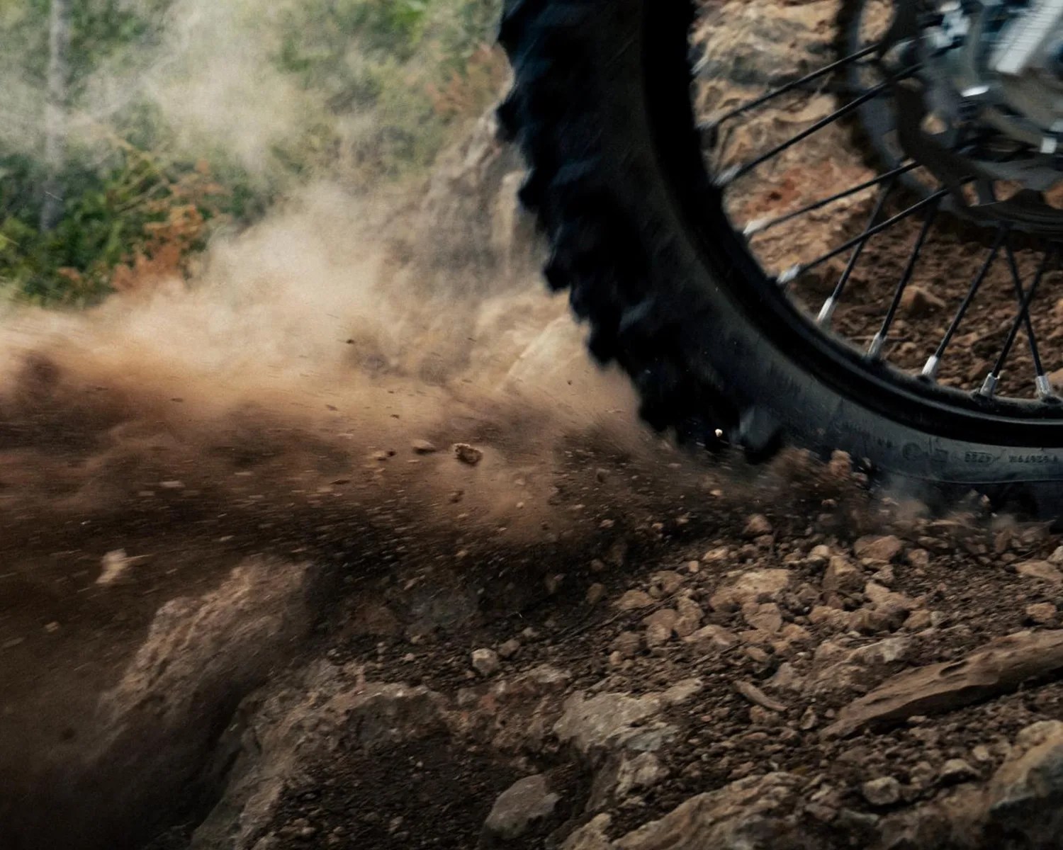 Close-up of a dirt bike tire kicking up dust and rocks on a rugged trail.