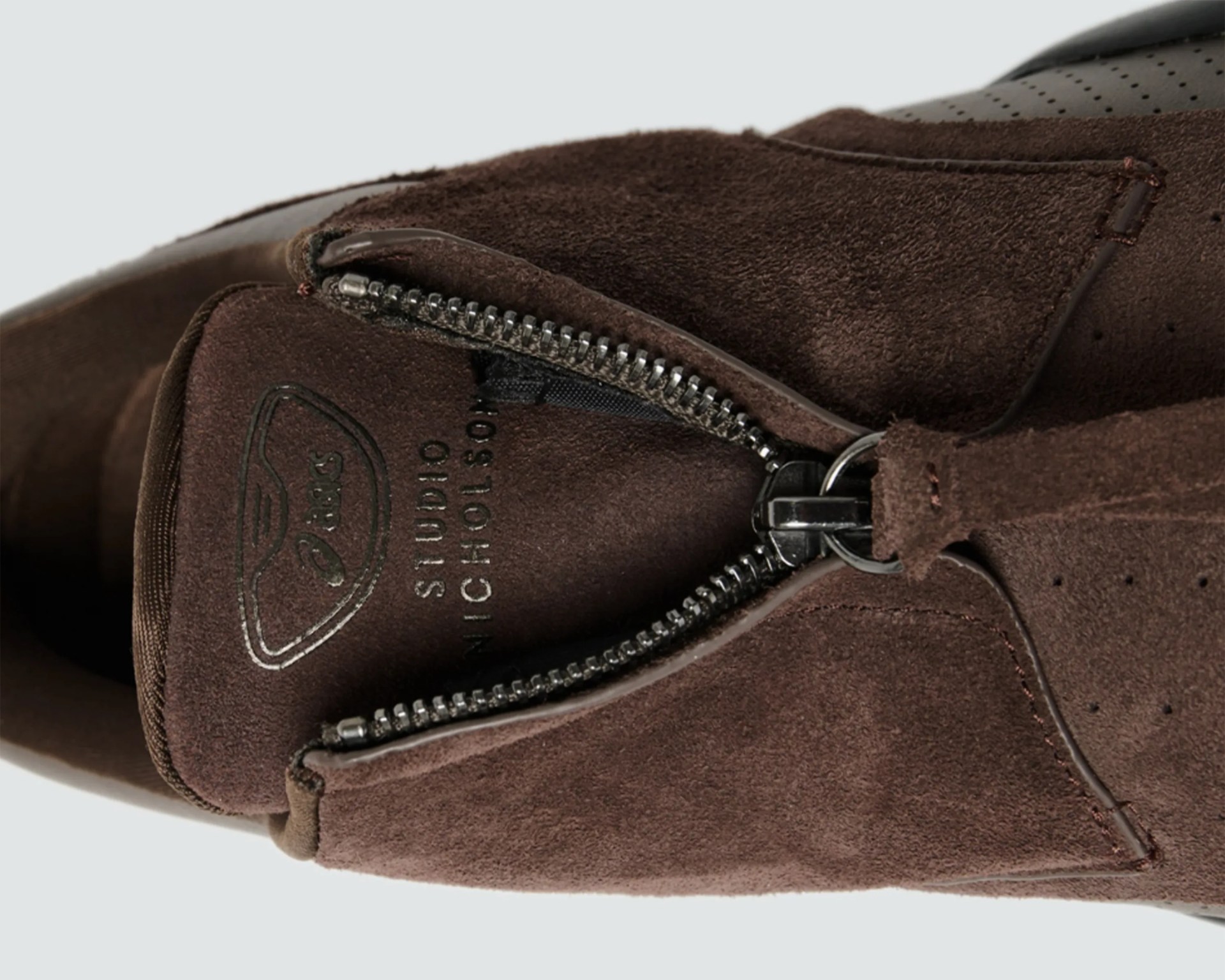 Close-up of a brown suede shoe with a partially open metal zipper and visible stitching.