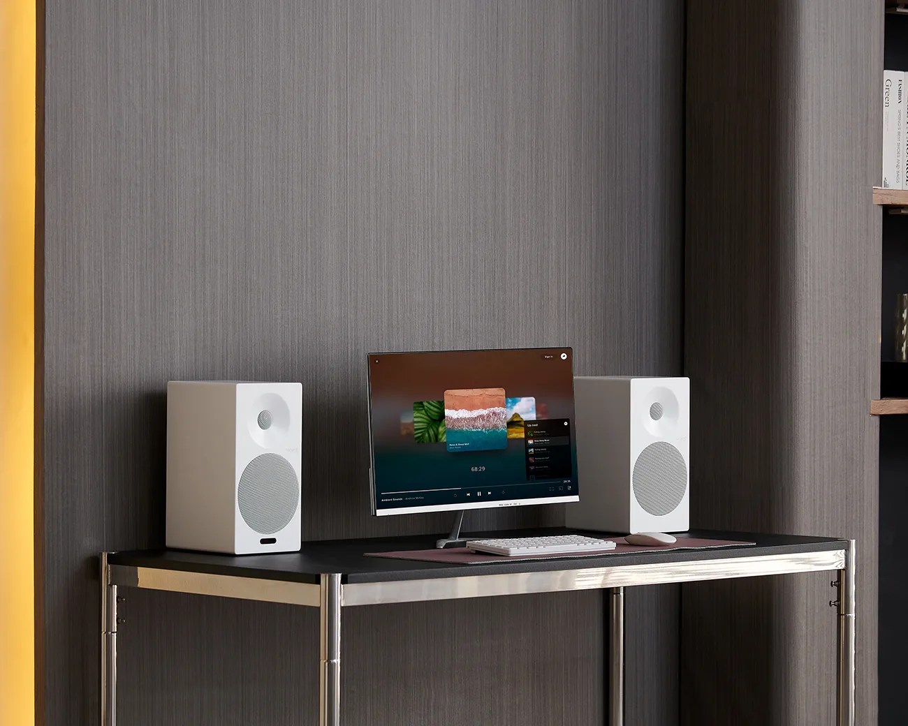 White desktop speakers flanking a slim monitor with a white keyboard and mouse on a black desk with metal legs.