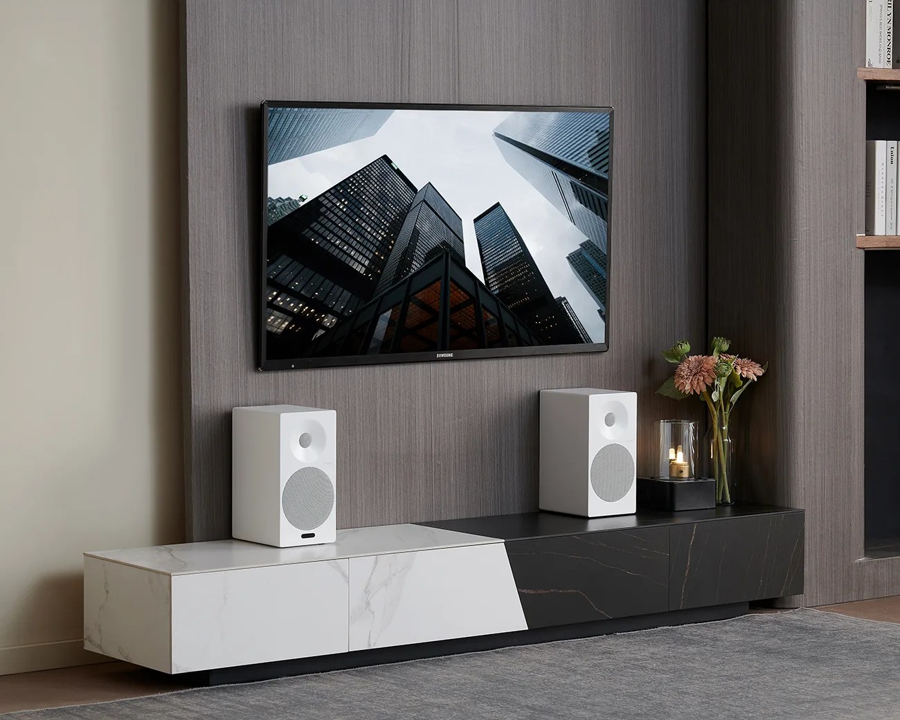 Wall-mounted flat-screen TV above a two-tone black and white marble media console with two white speakers and a vase with flowers.