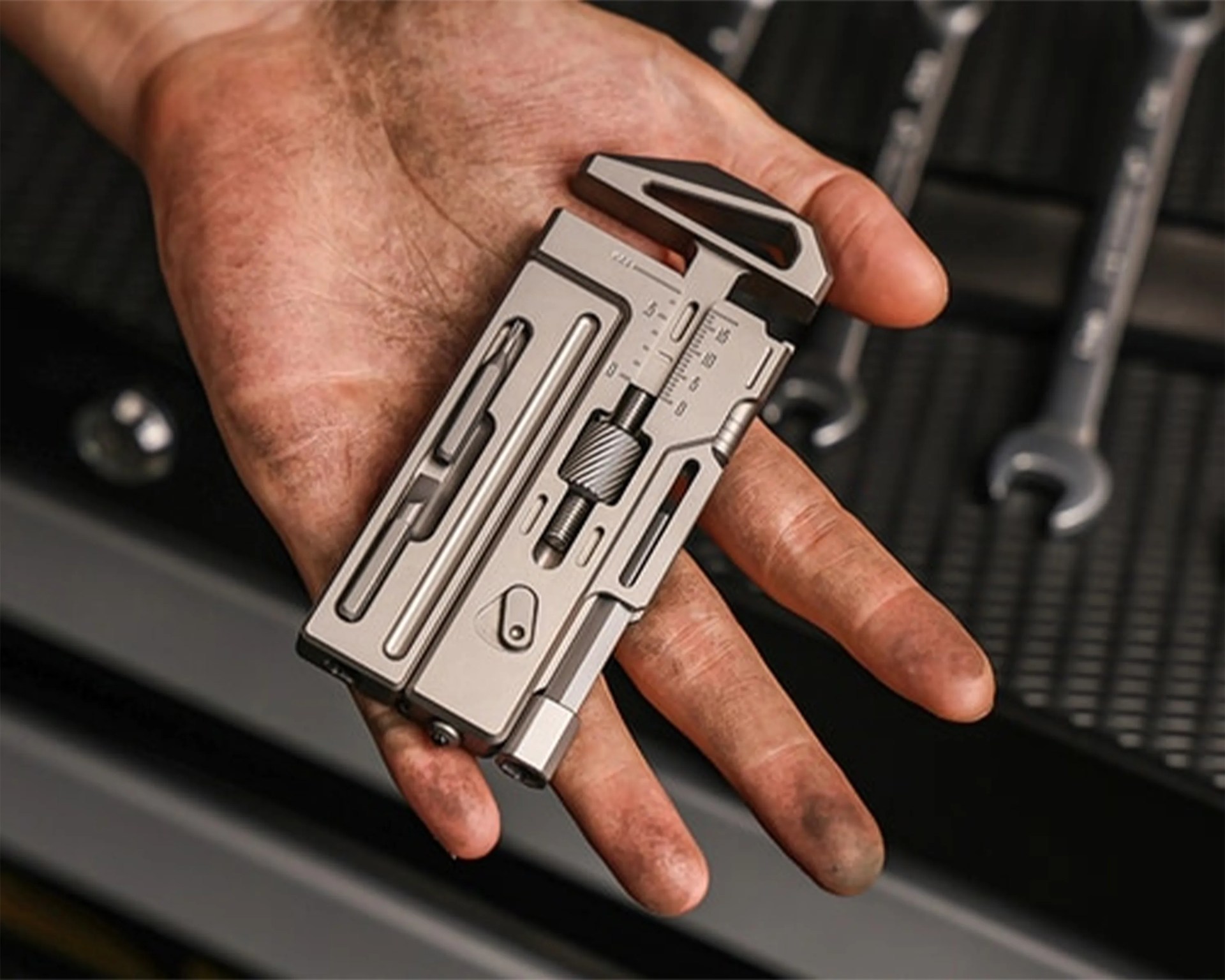 Compact silver multi-tool with various attachments held in a hand with a black tool mat and wrenches in the background.