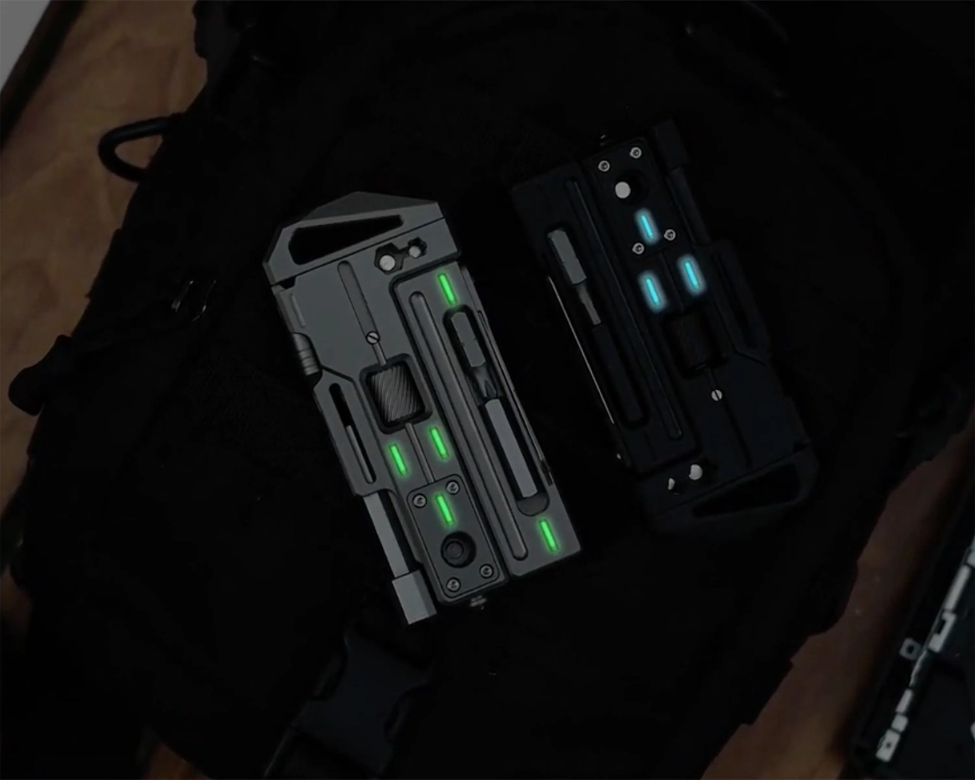 Two futuristic black and gray devices with glowing green and blue lights resting on a black fabric surface.