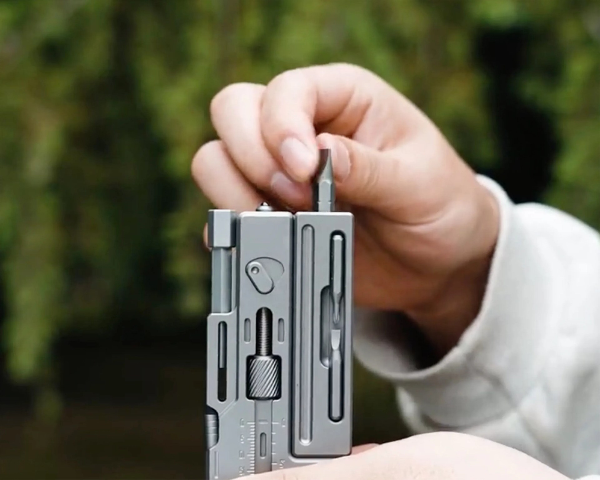 Hand holding a small gray multi-tool with a screwdriver bit being inserted.