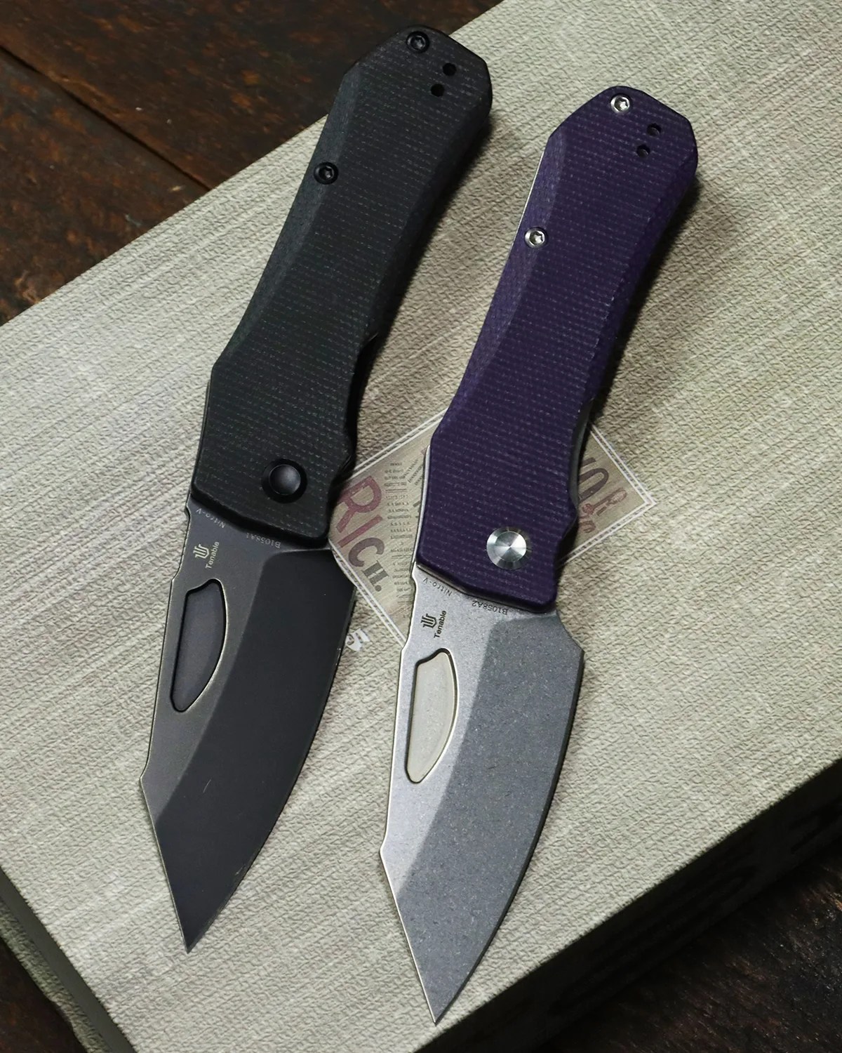 Two folding knives with textured handles, one black with a black blade and one purple with a silver blade, on a textured surface.