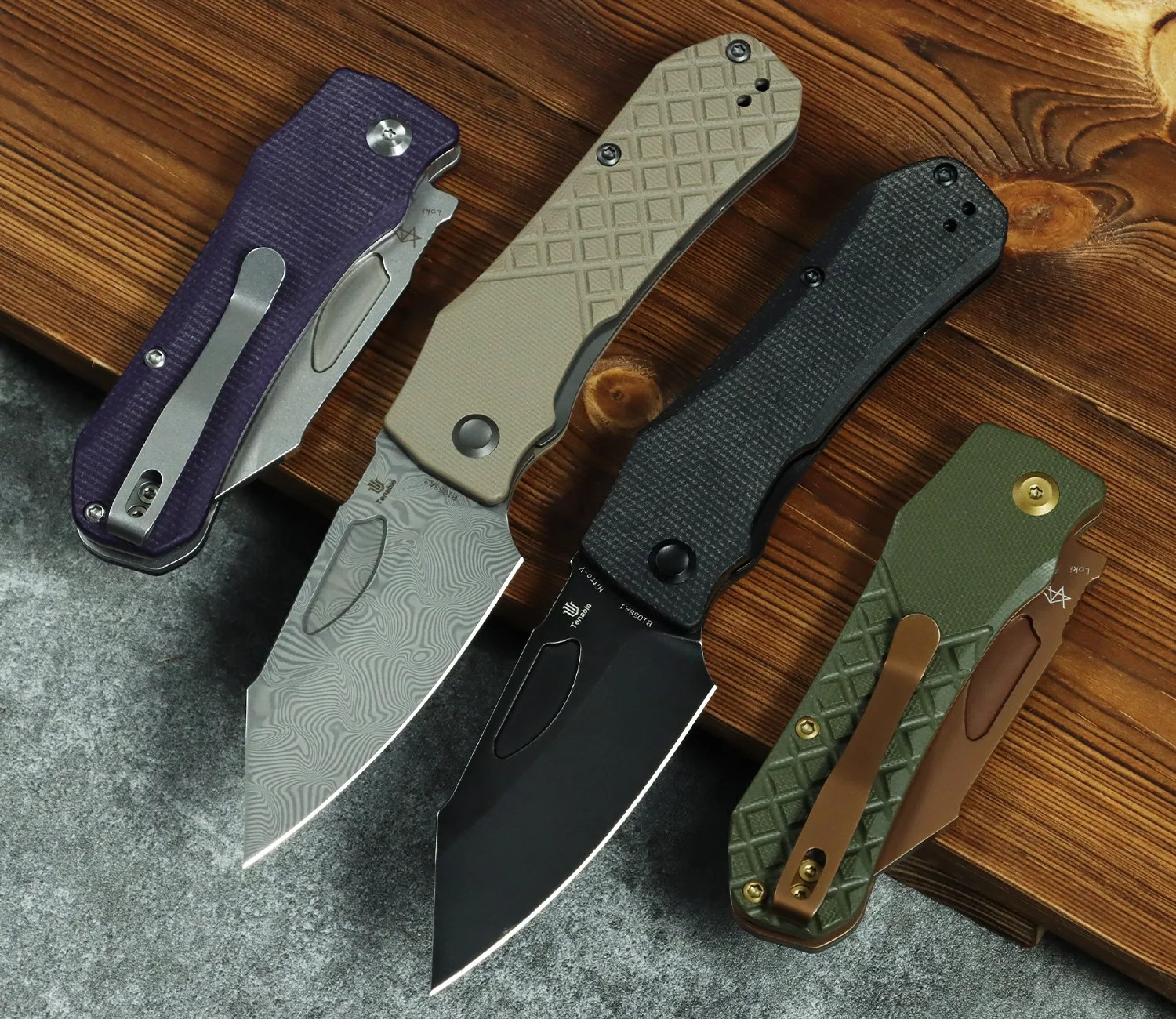 Four folding knives with textured handles in purple, tan, black, and green, two blades open showing patterned and black finishes, on wood and stone surfaces.