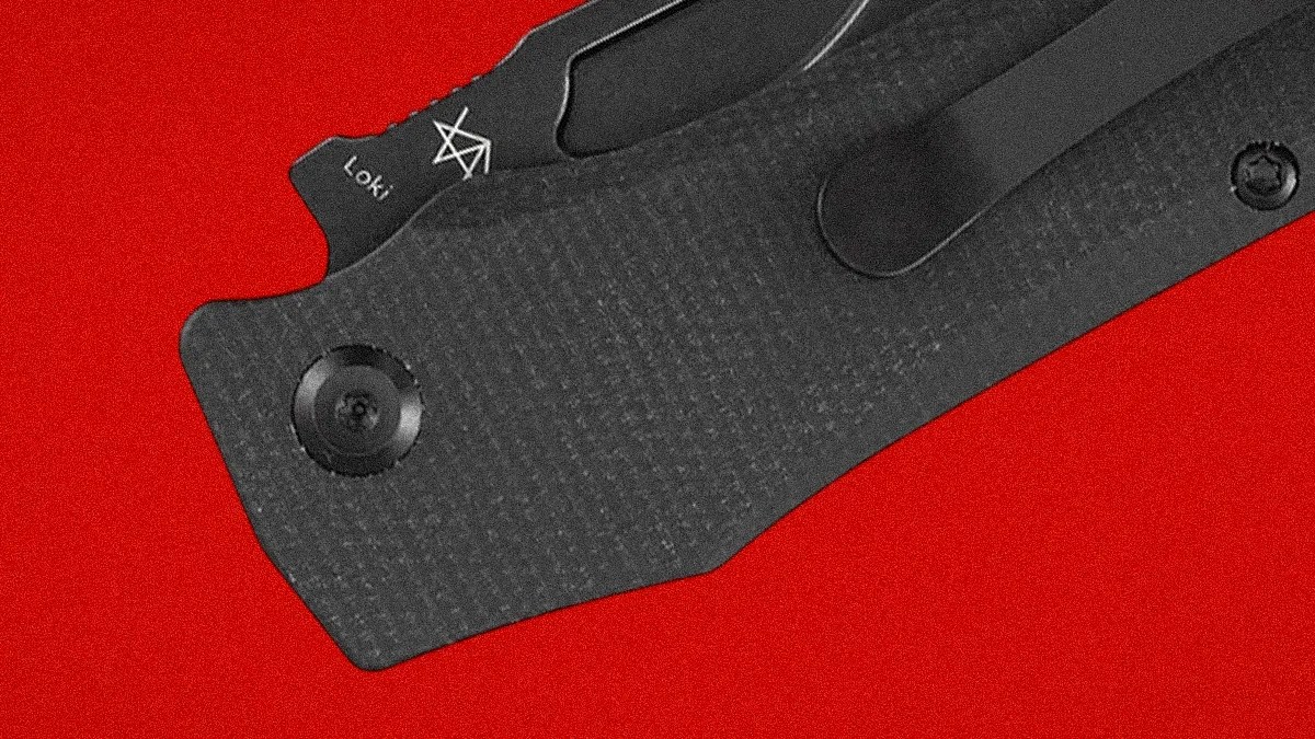 Close-up of a black textured folding knife handle with a clip and the word "Loki" on the blade against a red background.