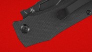 Close-up of a black textured folding knife handle with a clip and the word "Loki" on the blade against a red background.