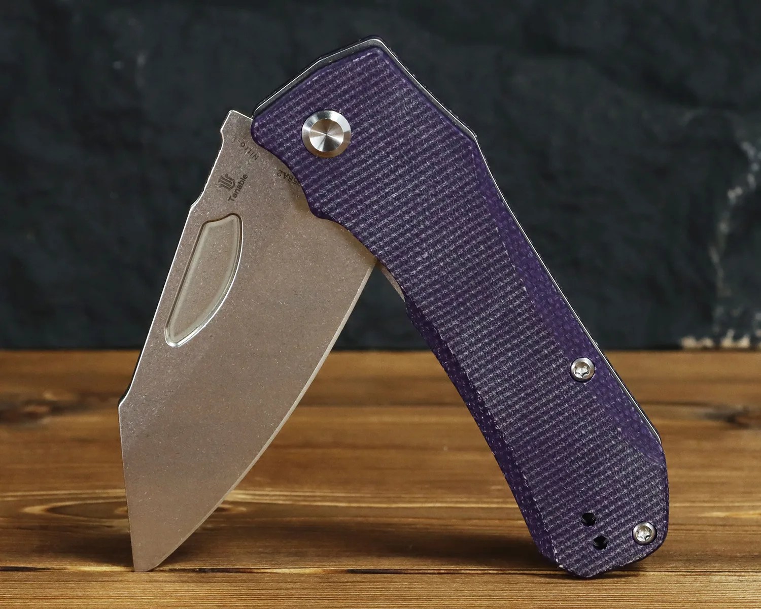 Folding knife with a stonewashed blade and textured purple handle standing open on a wooden surface.