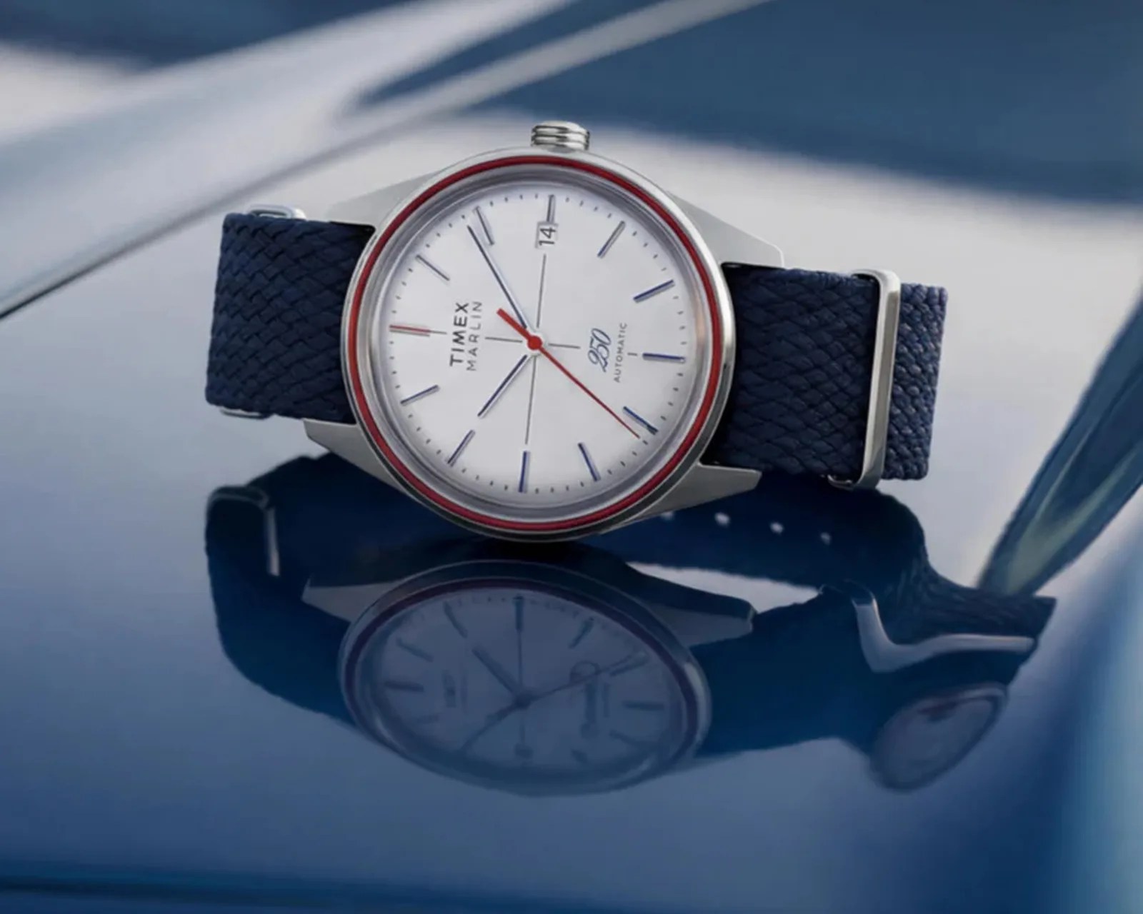 Silver Timex Marlin wristwatch with white dial, red second hand, and blue woven strap on reflective blue surface.
