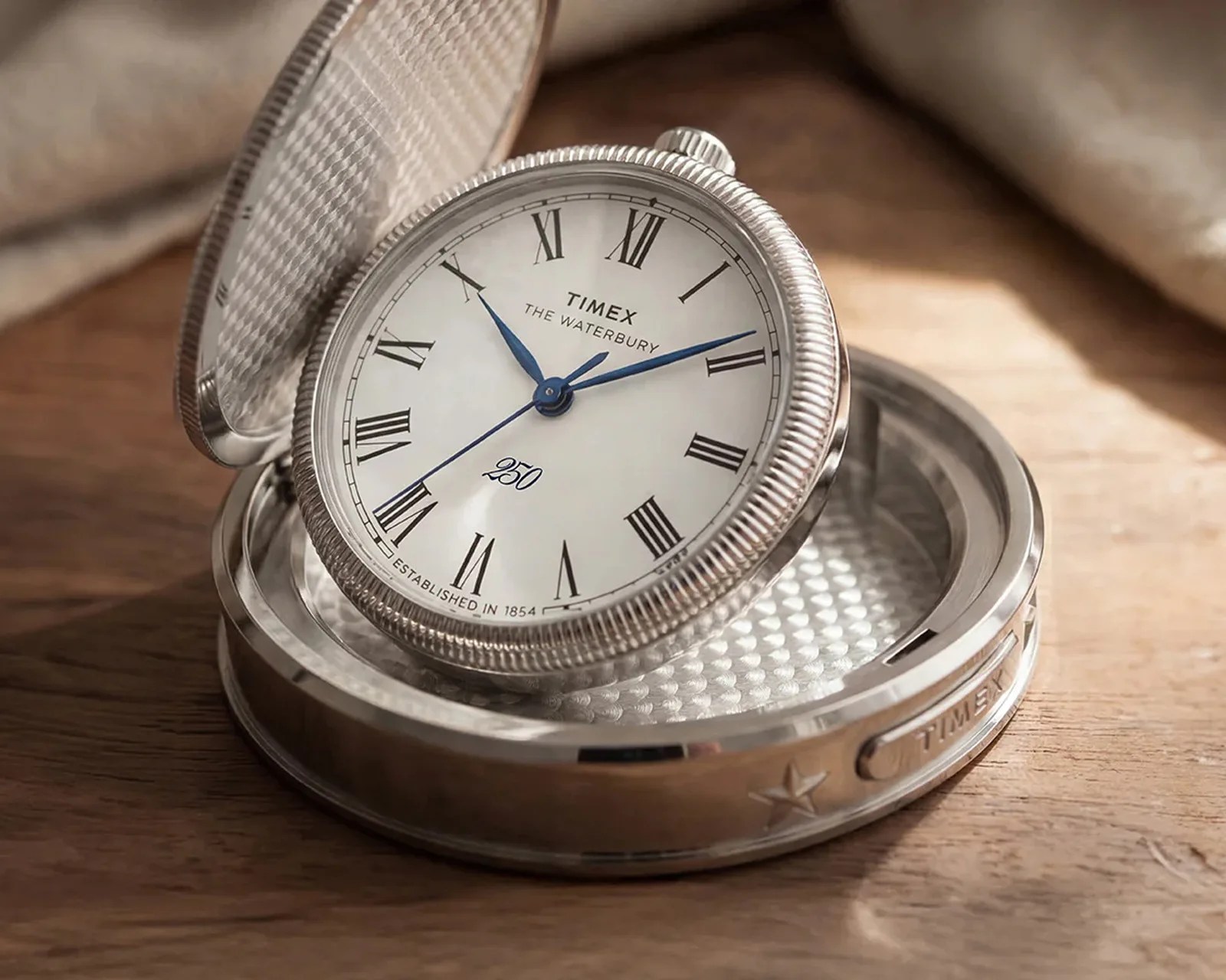 Silver Timex Waterbury table clock with Roman numerals and blue hands in an open case on a wooden surface.