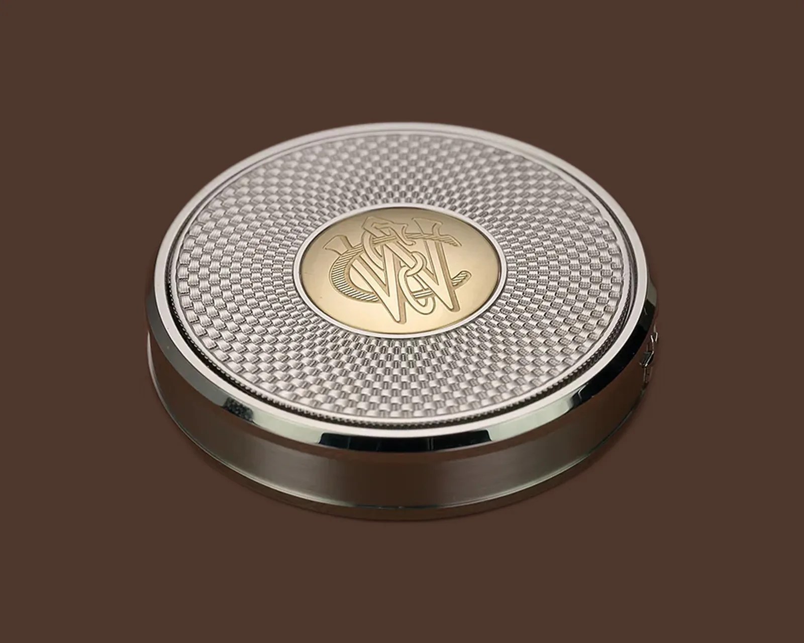 Round silver compact case with a textured pattern and a gold center medallion engraved with intertwined letters.