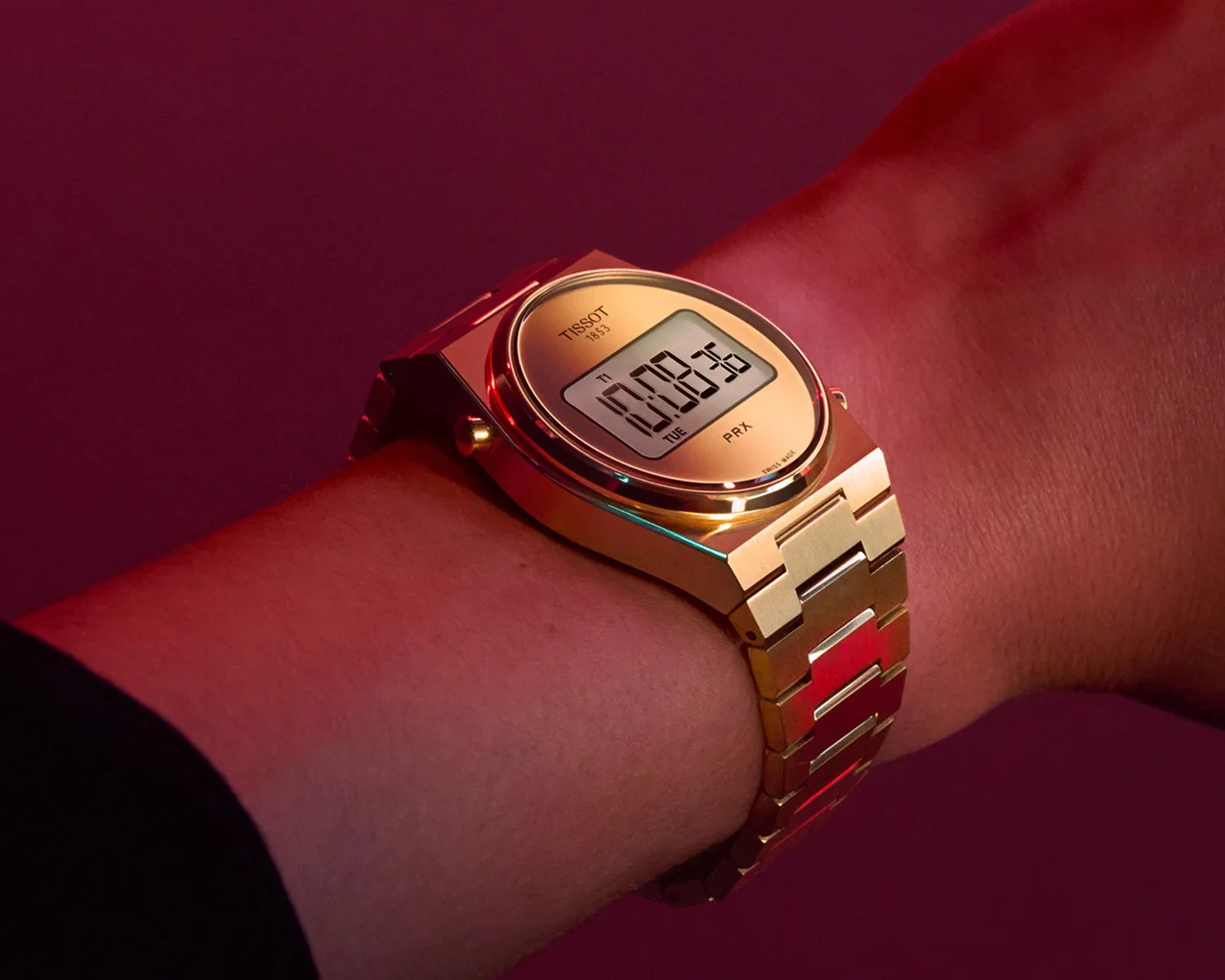 Gold-tone digital Tissot PRX watch with metal link bracelet on a wrist against a dark red background.