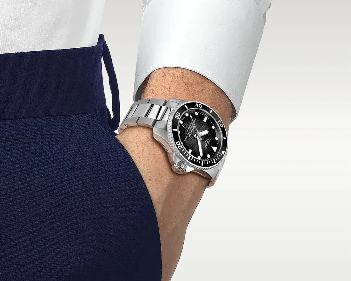 Silver metal wristwatch with black dial and rotating bezel worn on a wrist with a white shirt cuff and navy pants.