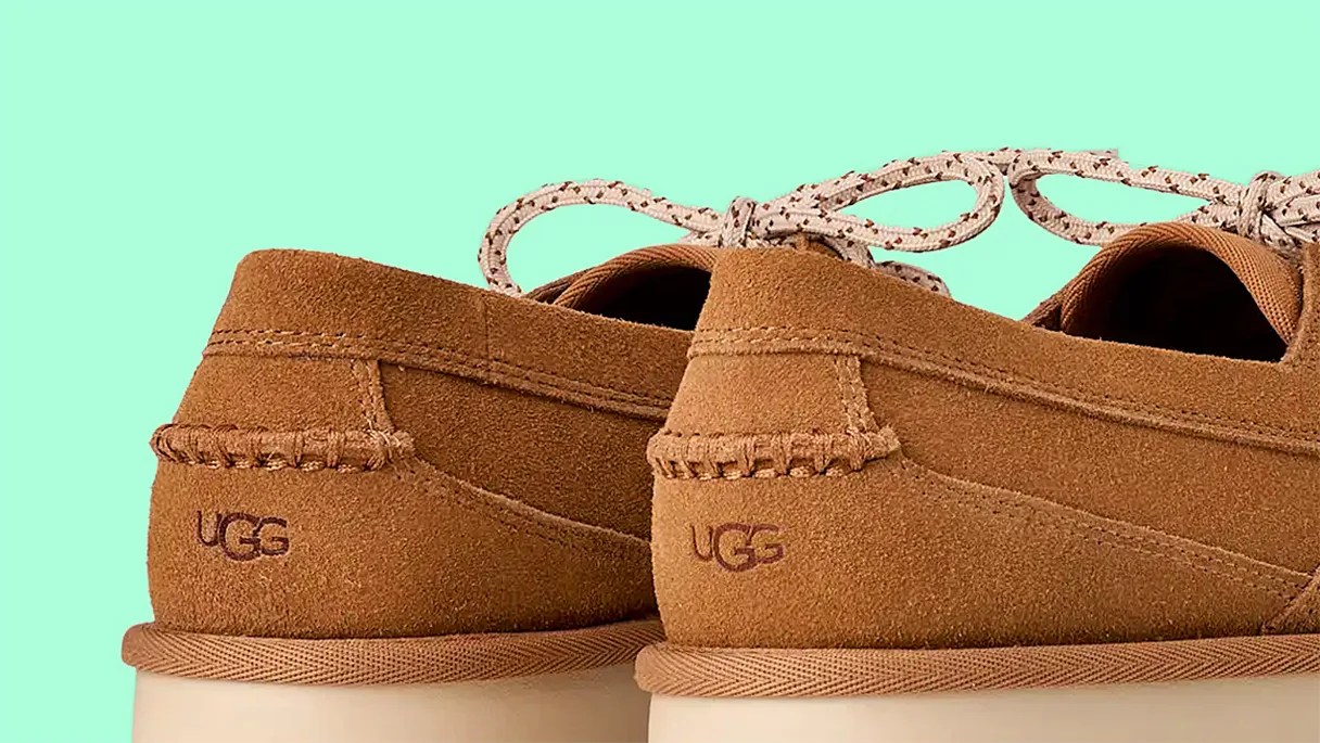 Pair of tan suede UGG shoes with stitched detailing and beige laces against a mint green background.