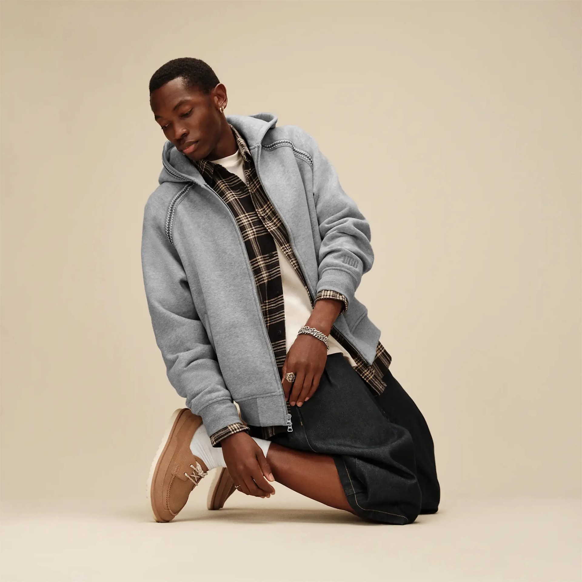 Person wearing a light gray zip-up hoodie over a black and beige plaid shirt, black skirt, white socks, and tan sneakers kneeling on a beige floor.