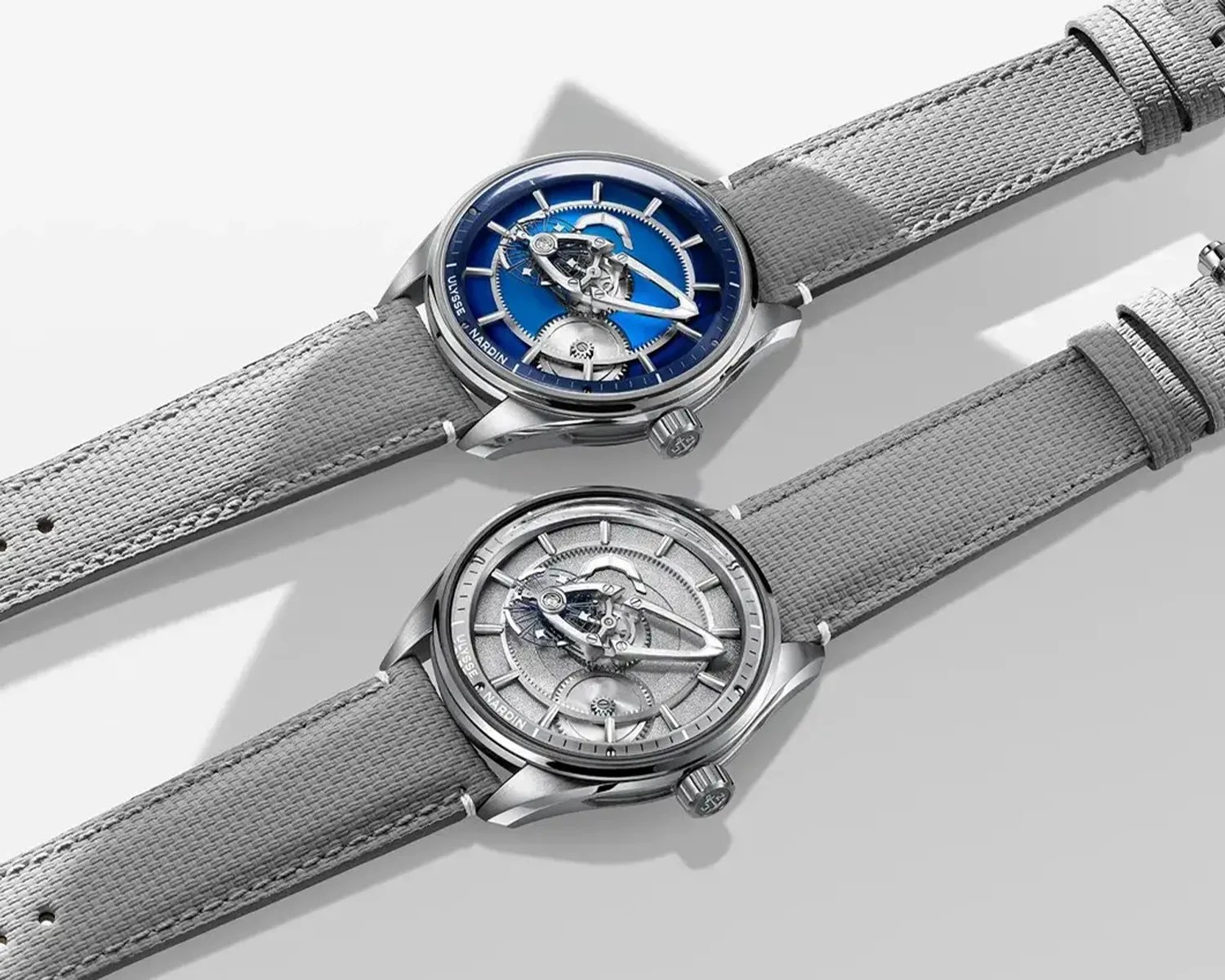 Two Ulysse Nardin wristwatches with silver cases and gray fabric straps, one with a blue dial and the other with a silver dial.