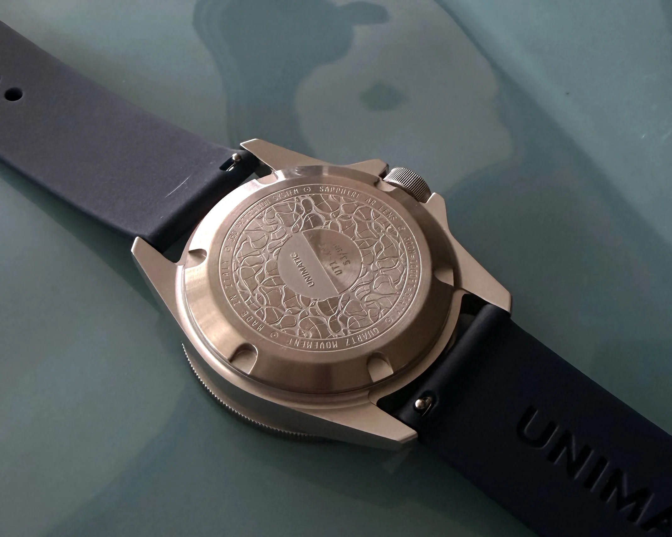Back of a bronze wristwatch with engraved design and black rubber strap on a glass surface.