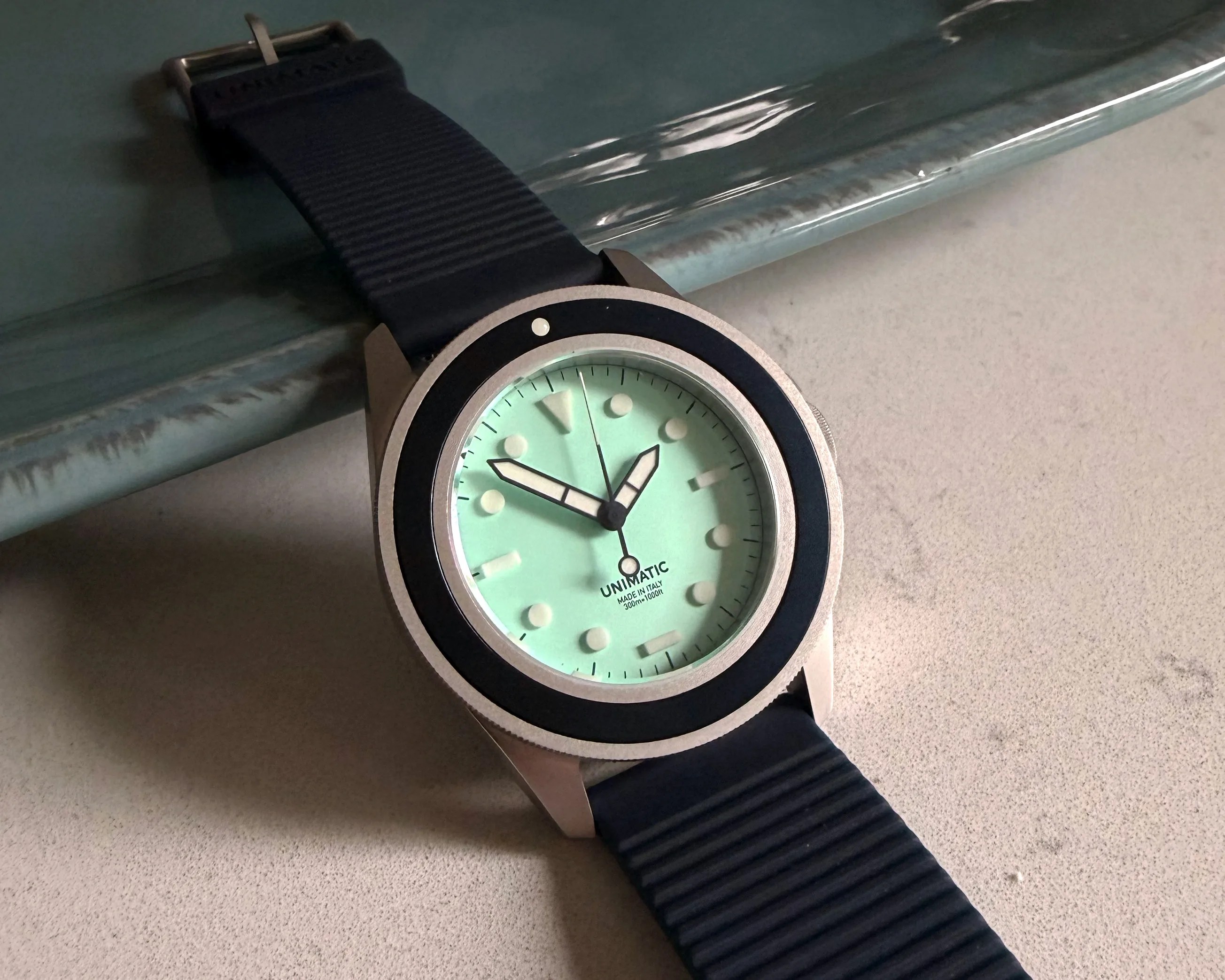Watch with a mint green dial, black bezel, and black ribbed strap on a light surface.