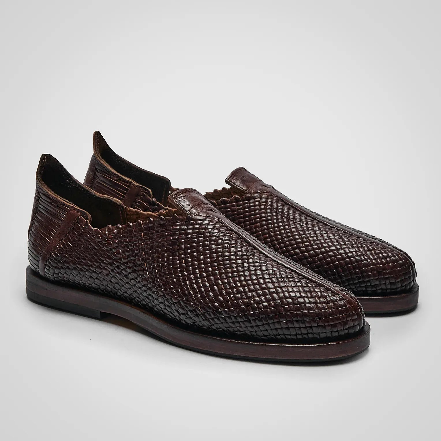 Pair of dark brown woven leather slip-on shoes with scalloped edges and low wooden soles.