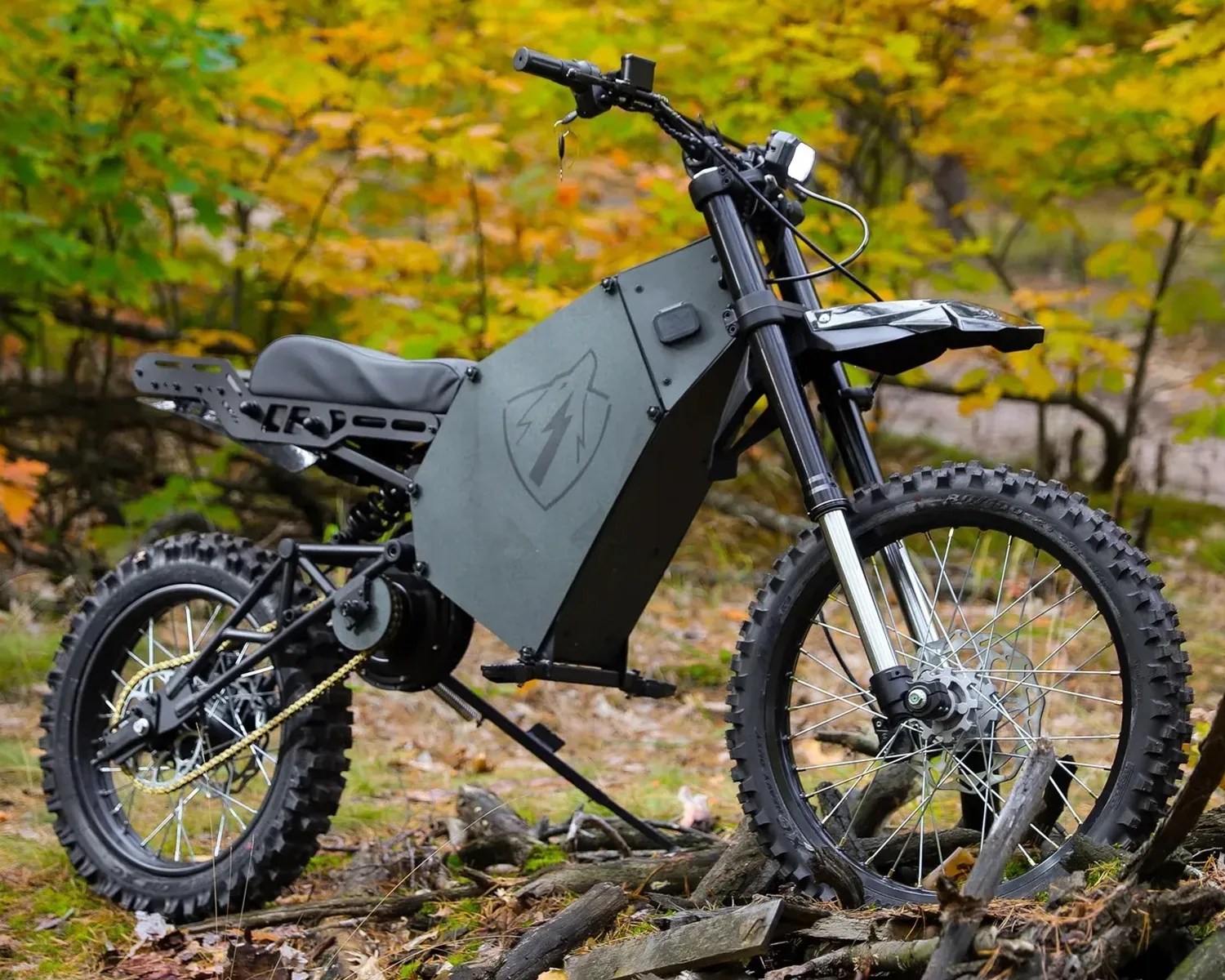 Black electric dirt bike with knobby tires and angular battery compartment parked on forest ground.