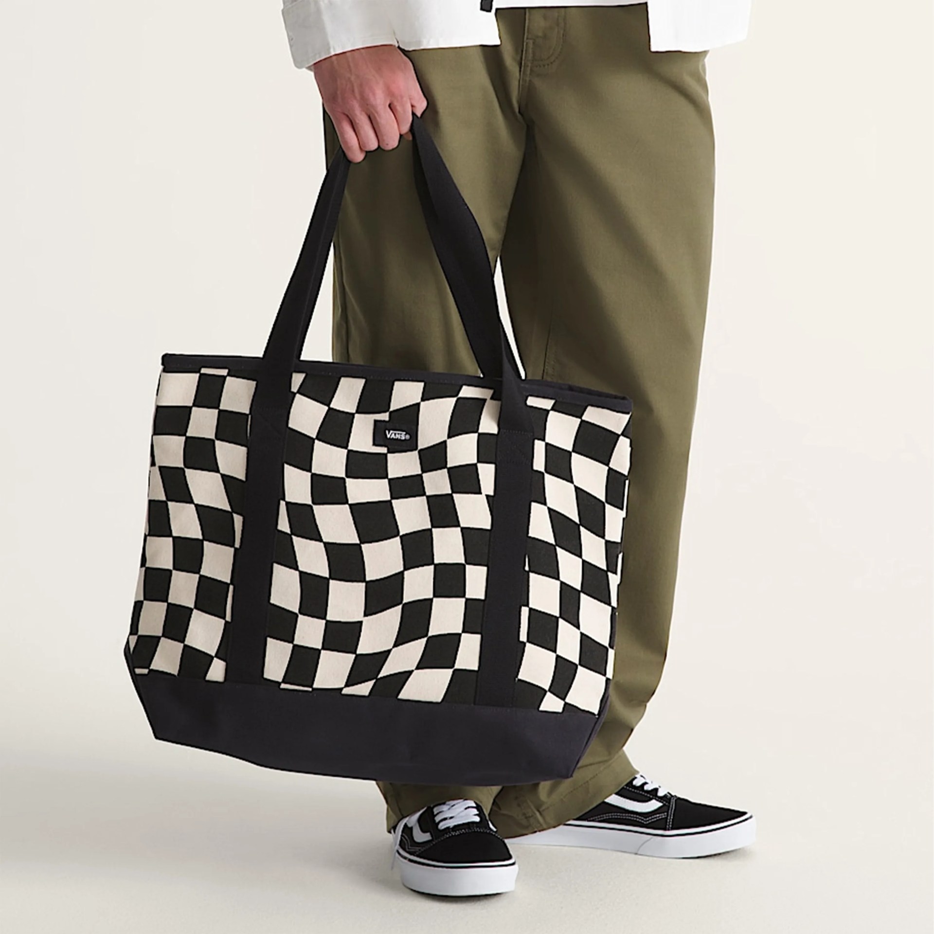 Black and white checkered Vans tote bag held by a person wearing olive pants and black Vans sneakers.