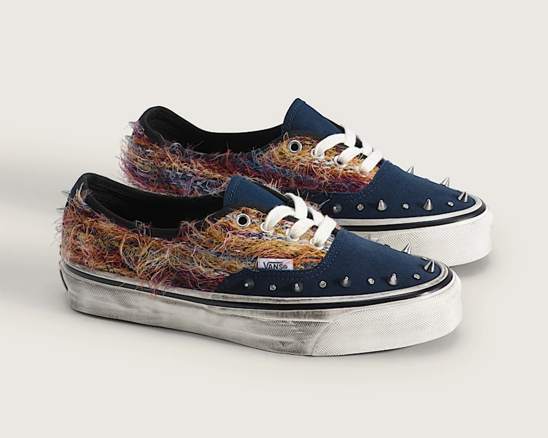Pair of Vans sneakers with navy blue toe caps, multicolored fuzzy fabric sides, white laces, and silver spikes on the toe area.