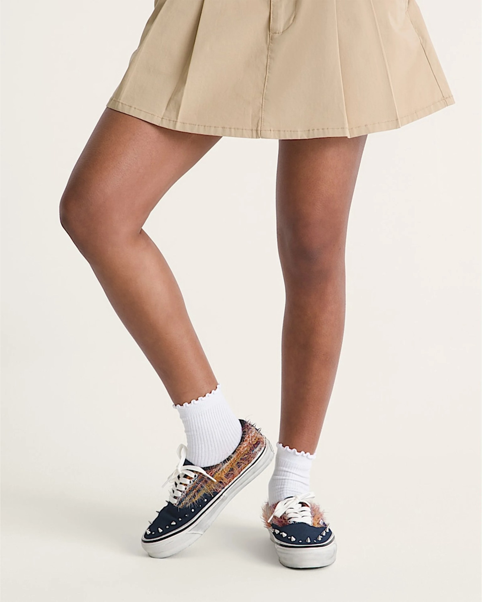 Legs wearing a beige pleated skirt, white ruffled ankle socks, and navy sneakers with multicolored fuzzy accents and silver spikes.