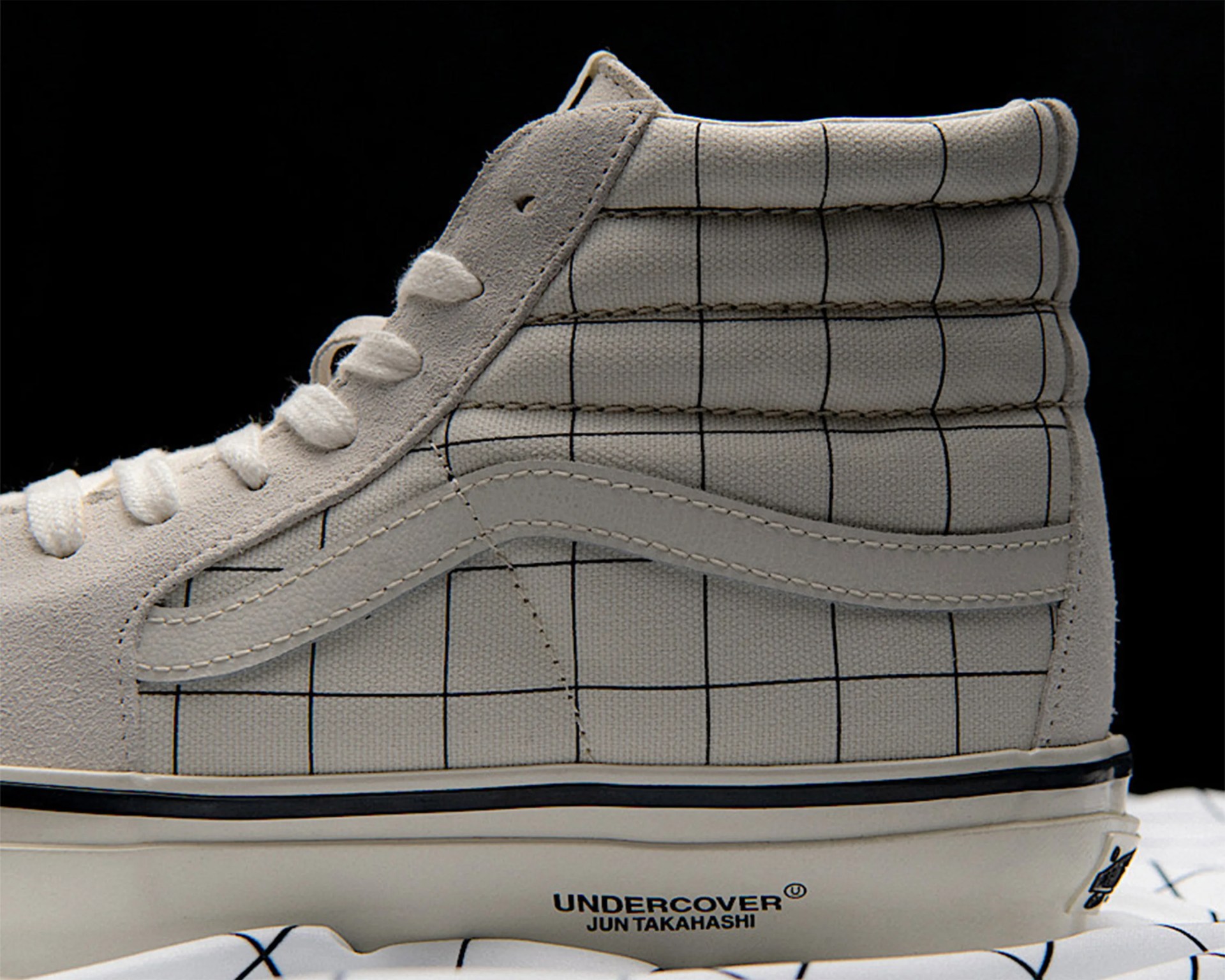 High-top sneaker with beige suede and grid-patterned fabric upper, "UNDERCOVER JUN TAKAHASHI" text on the white midsole.
