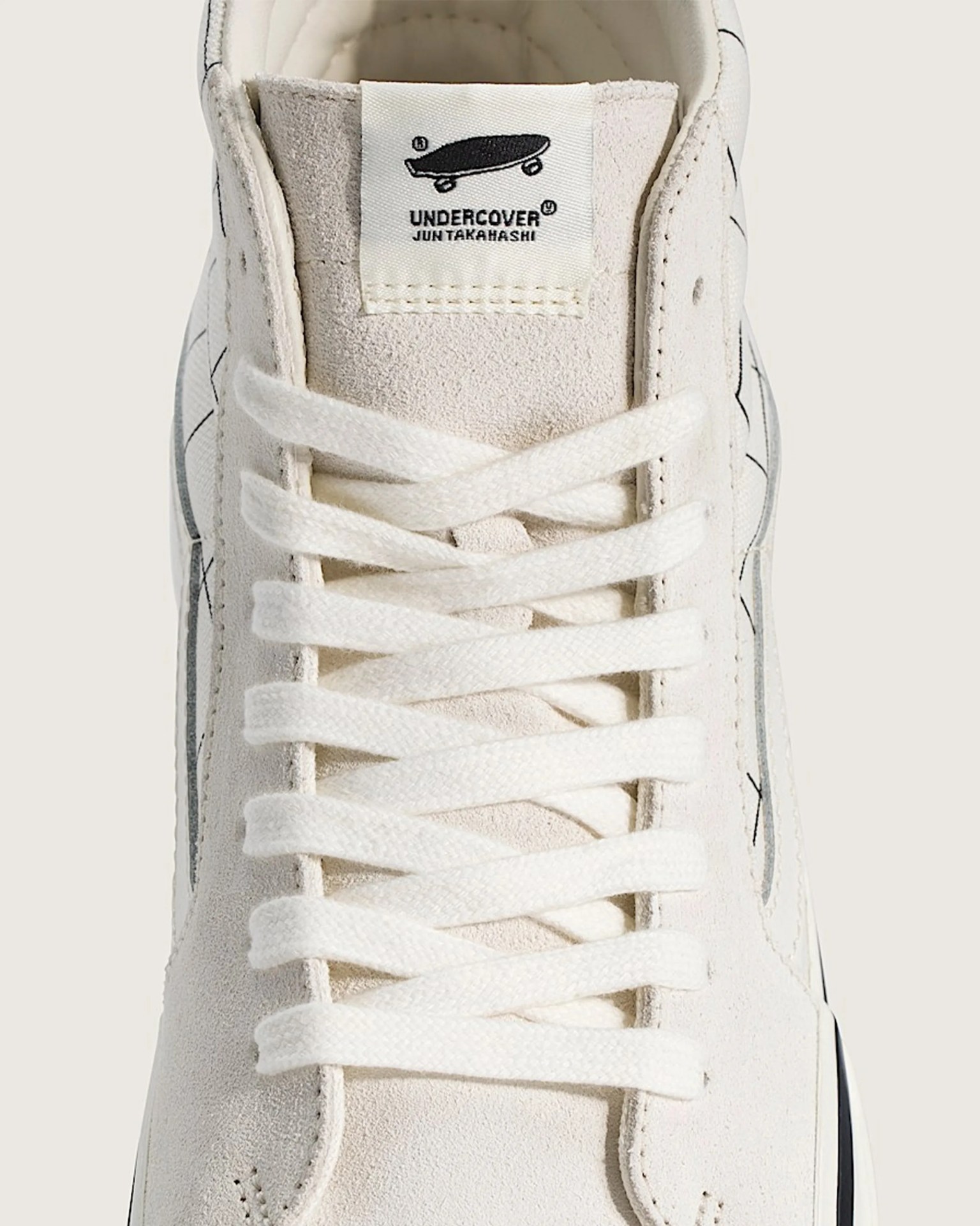 Close-up of cream suede sneaker tongue with white label showing black skateboard logo and "UNDERCOVER JUN TAKAHASHI" text.