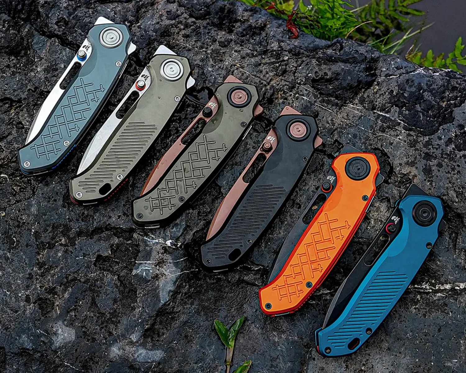Six folding knives with partially visible blades in various colors including blue, olive, bronze, black, orange, and teal, arranged on a dark rocky surface.