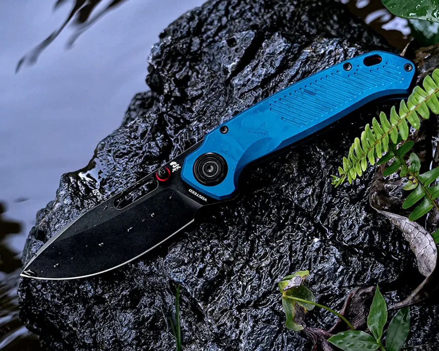 Black folding knife with a blue textured handle resting on wet dark rock near green foliage.